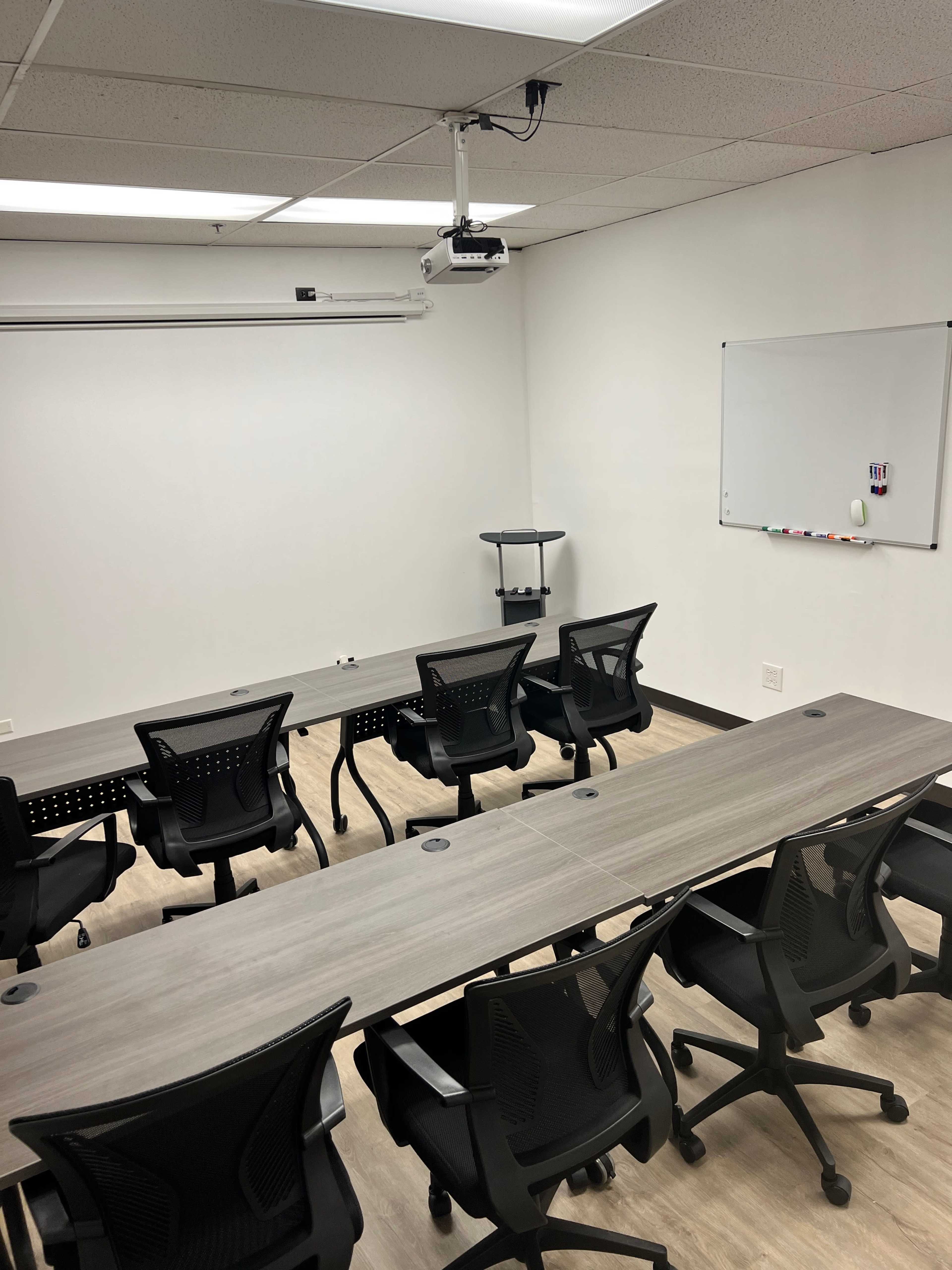 A modern conference room features a long table with multiple black chairs, a projector on the ceiling, and a whiteboard on the wall.