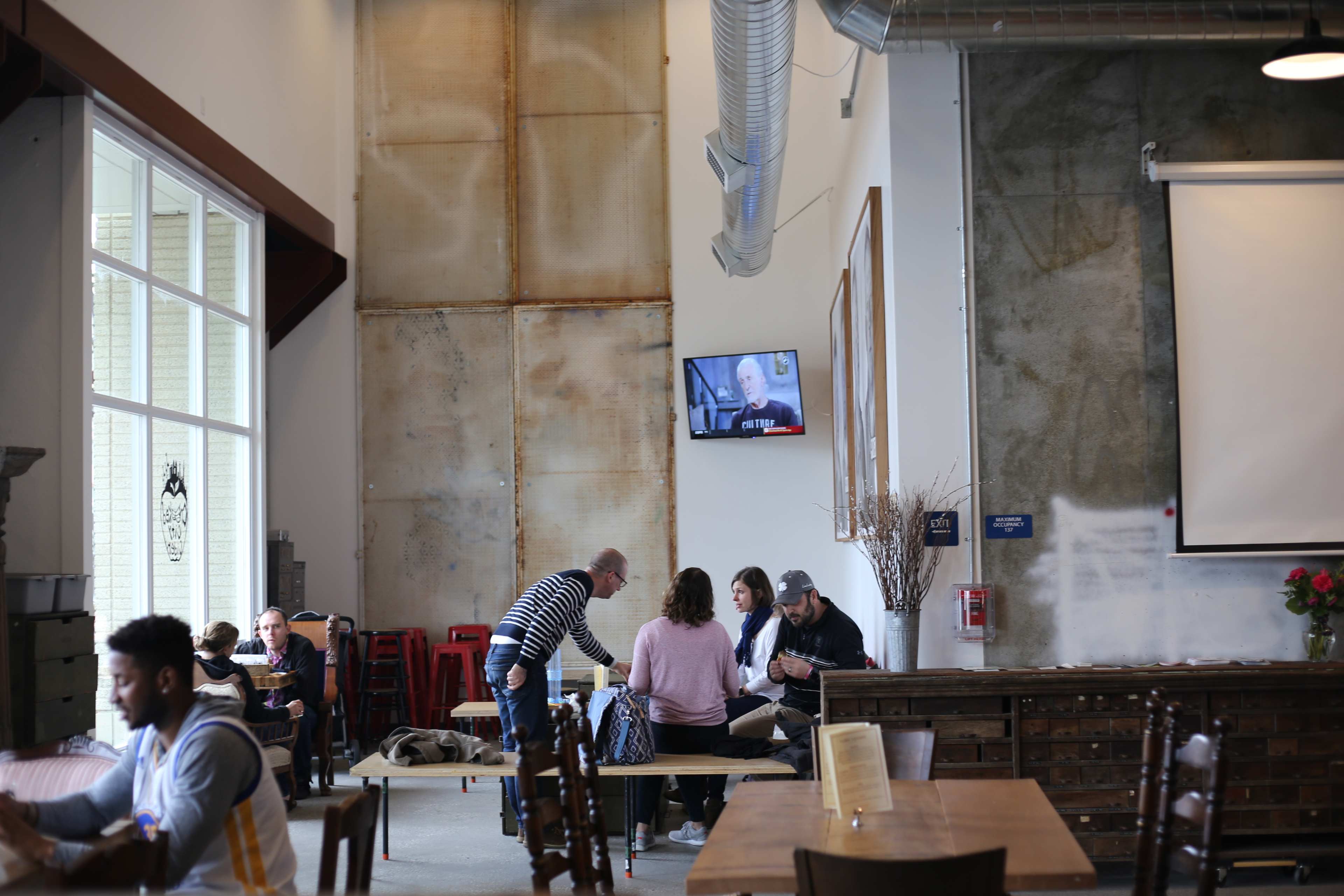 The interior of a café features a mix of tables and chairs, with patrons engaged in conversation and a television screen displaying a program on the wall.