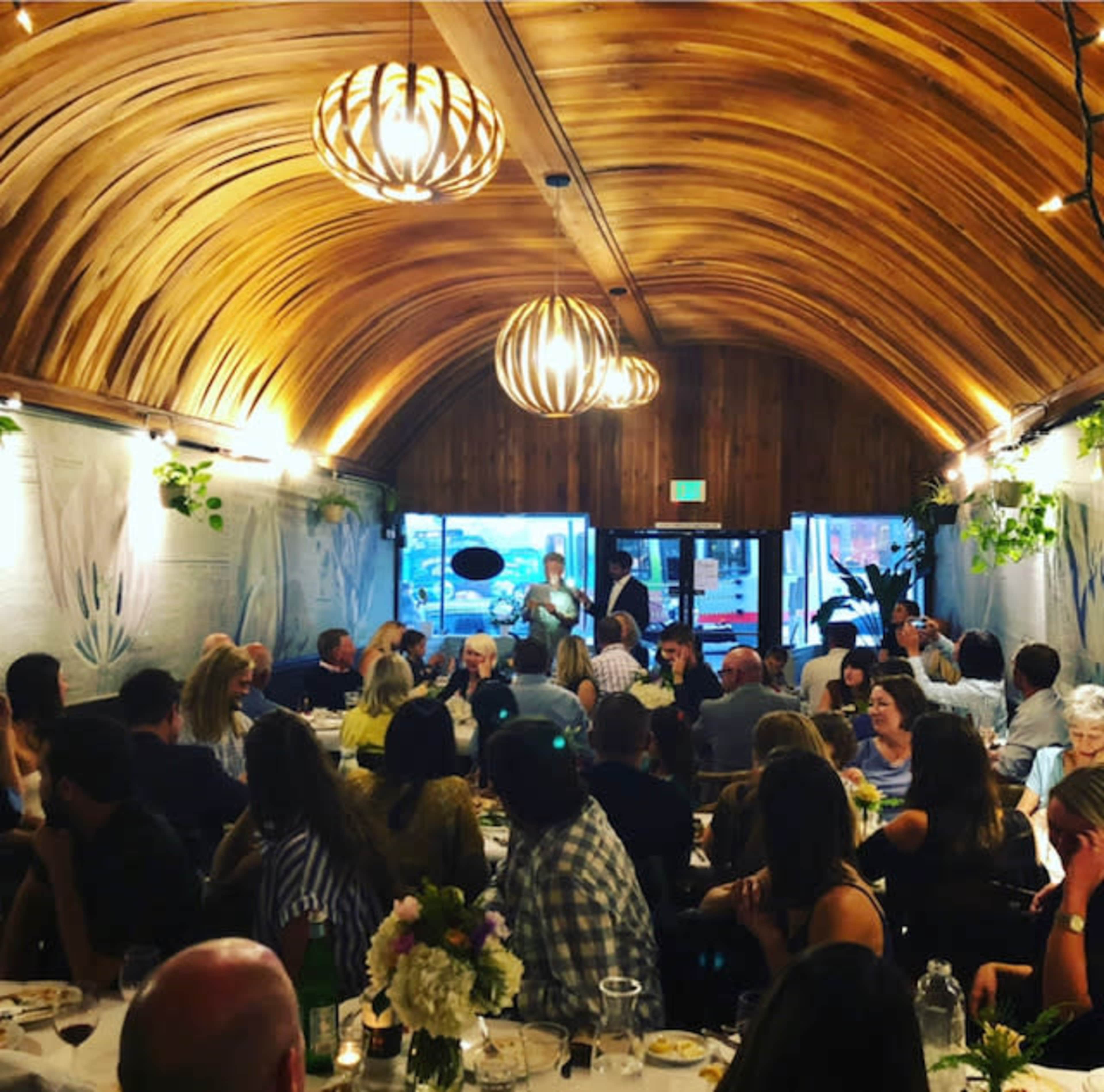 A crowded restaurant features a wooden, curved ceiling with pendant lights, as guests dine at tables adorned with flowers.