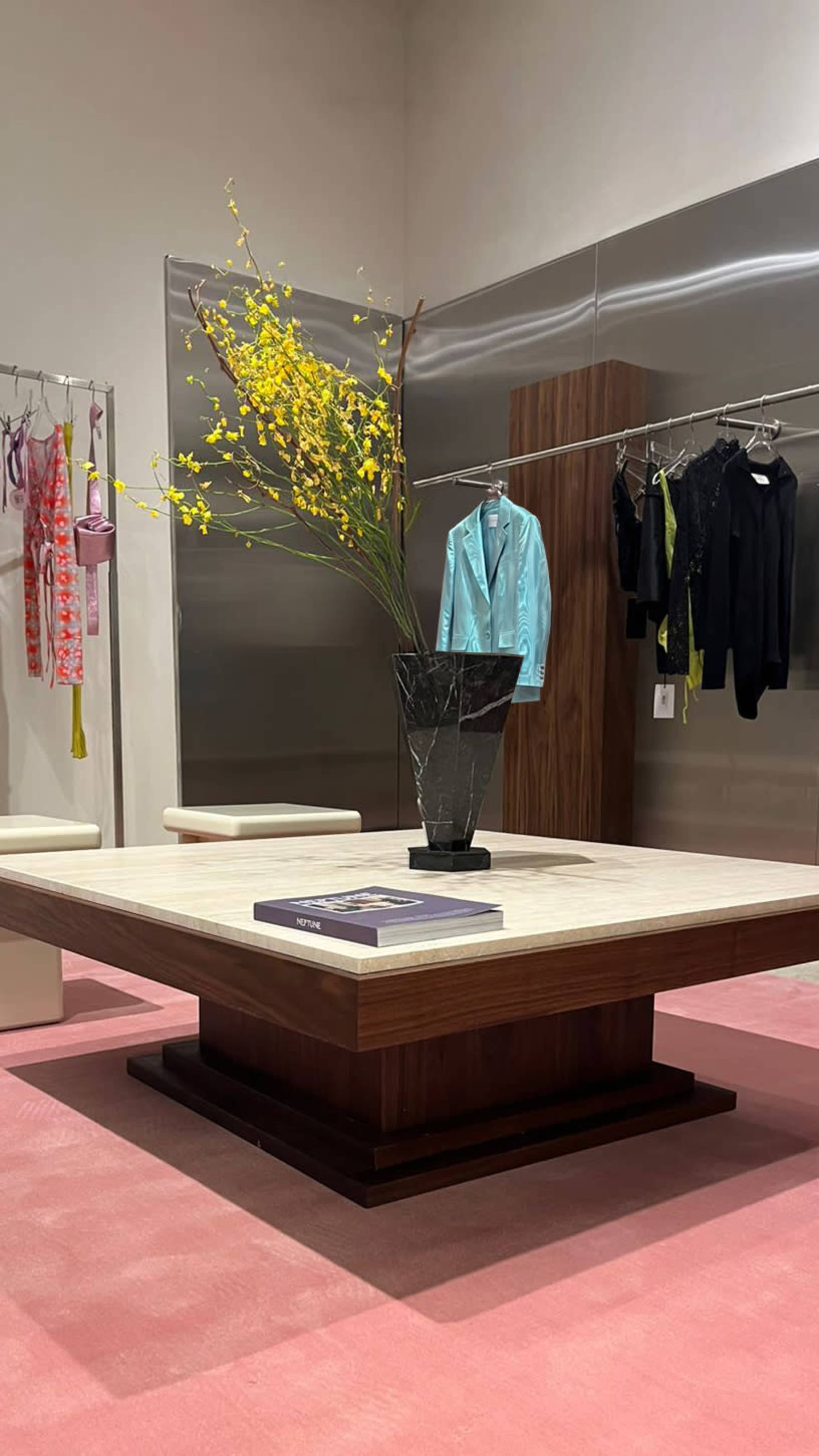 The image depicts a modern retail display featuring a large square table with a decorative vase of yellow flowers, surrounded by clothing hung on racks in a minimalist space.