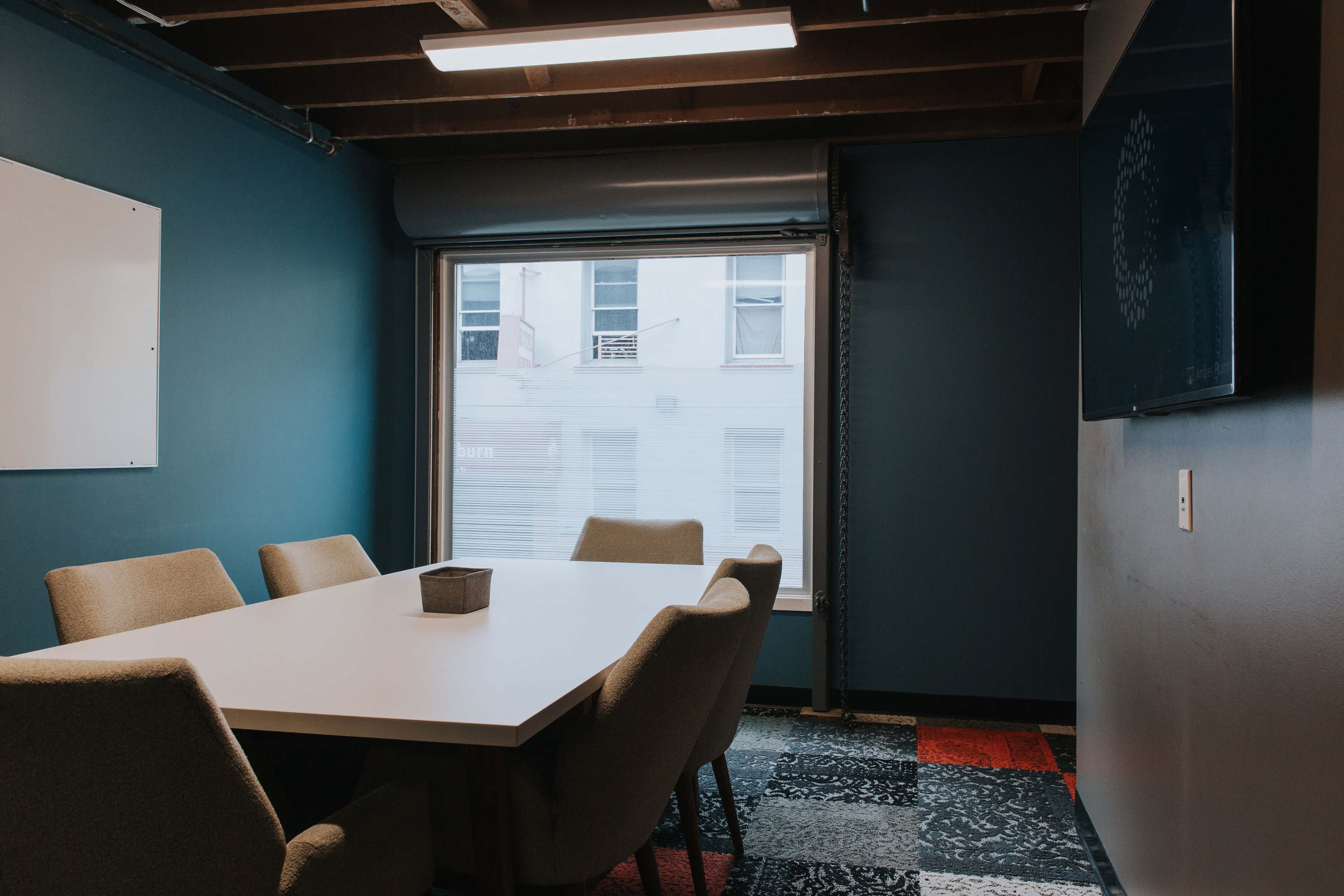 A meeting room features a rectangular white table surrounded by beige chairs, with a window showing a view of the building outside.