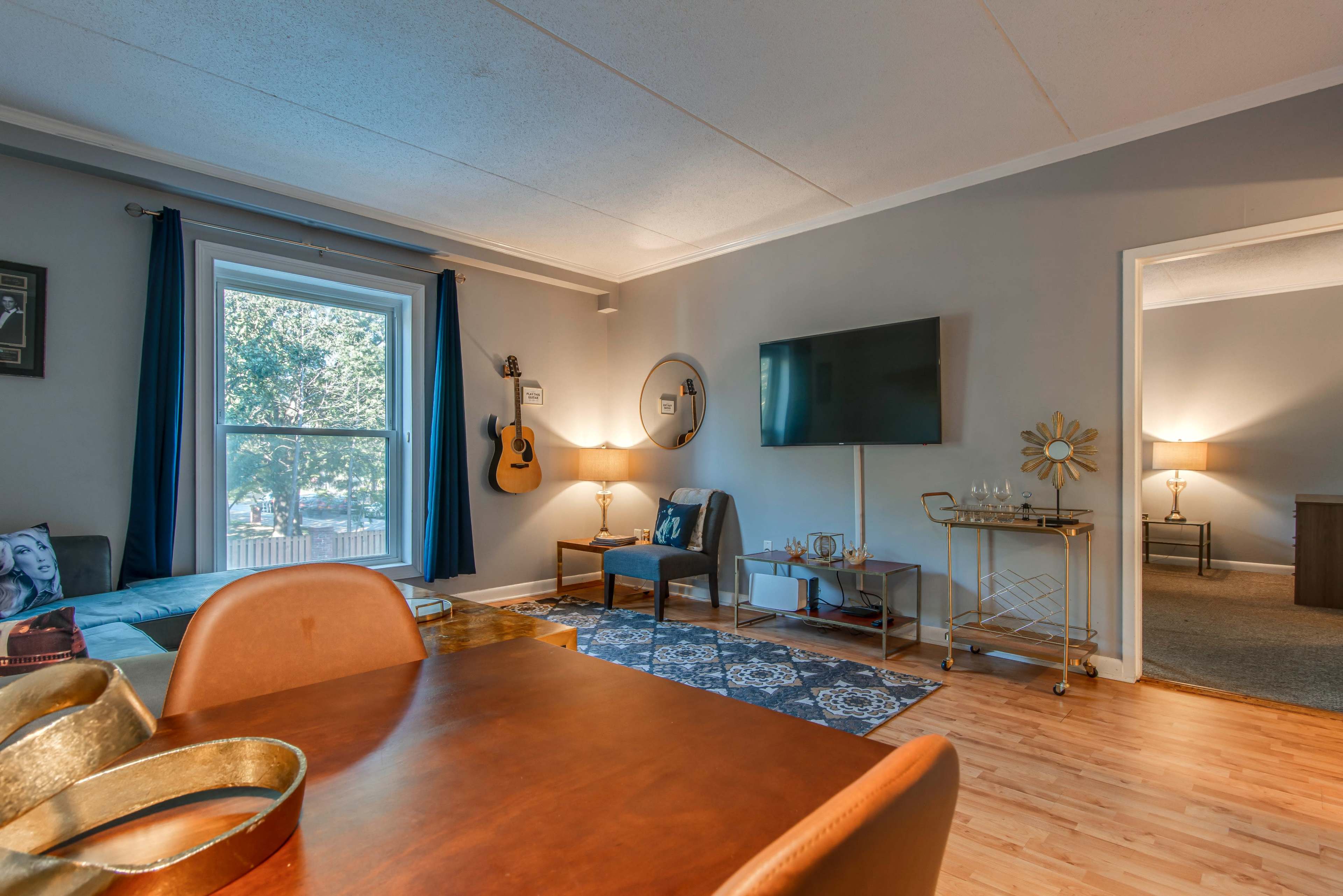 The living room features a guitar on the wall, a television mounted on the opposite wall, and a wooden dining table in the foreground.