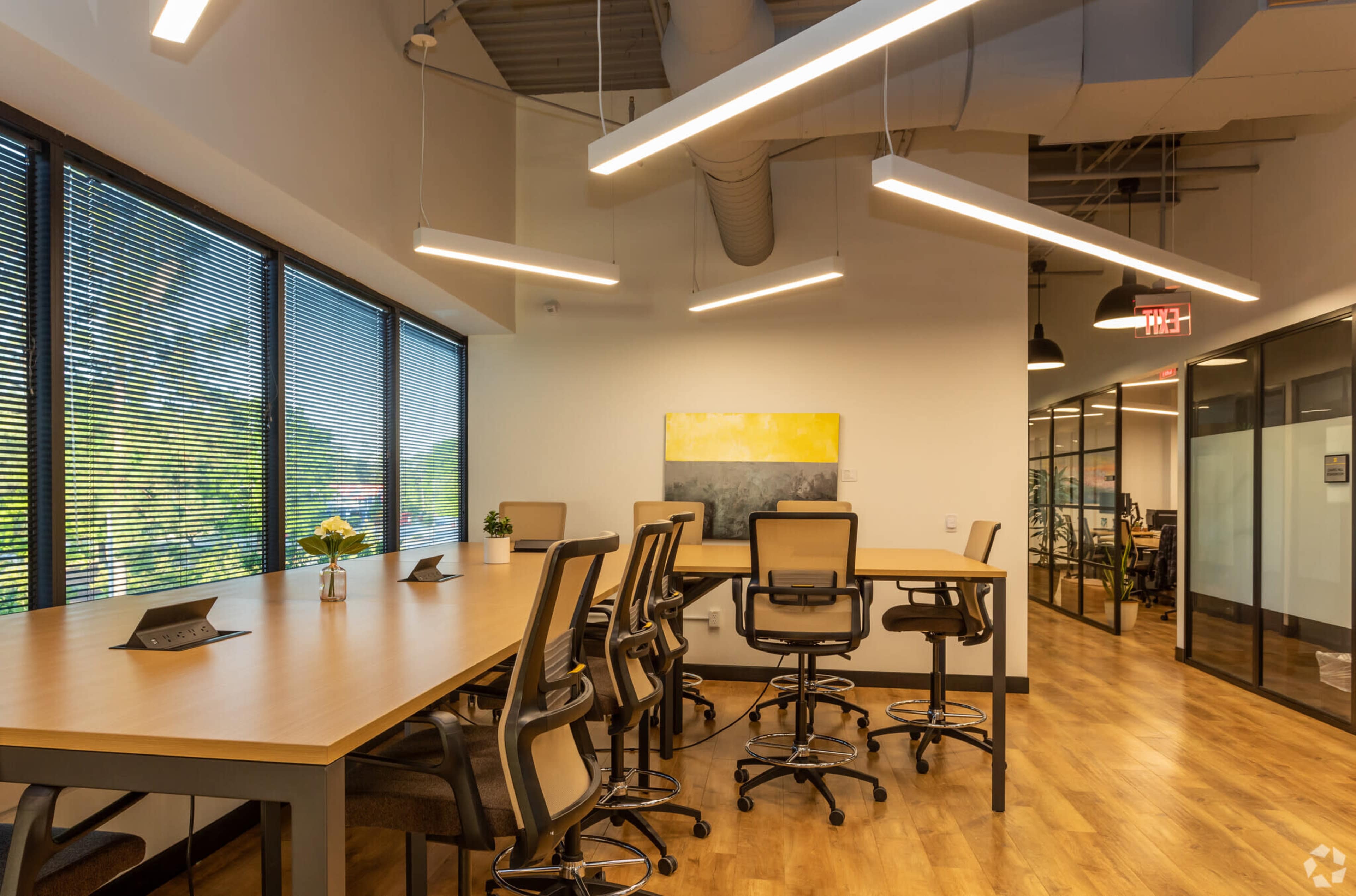 The image shows a modern office conference room with large windows, a long wooden table surrounded by chairs, and framed artwork on the wall.