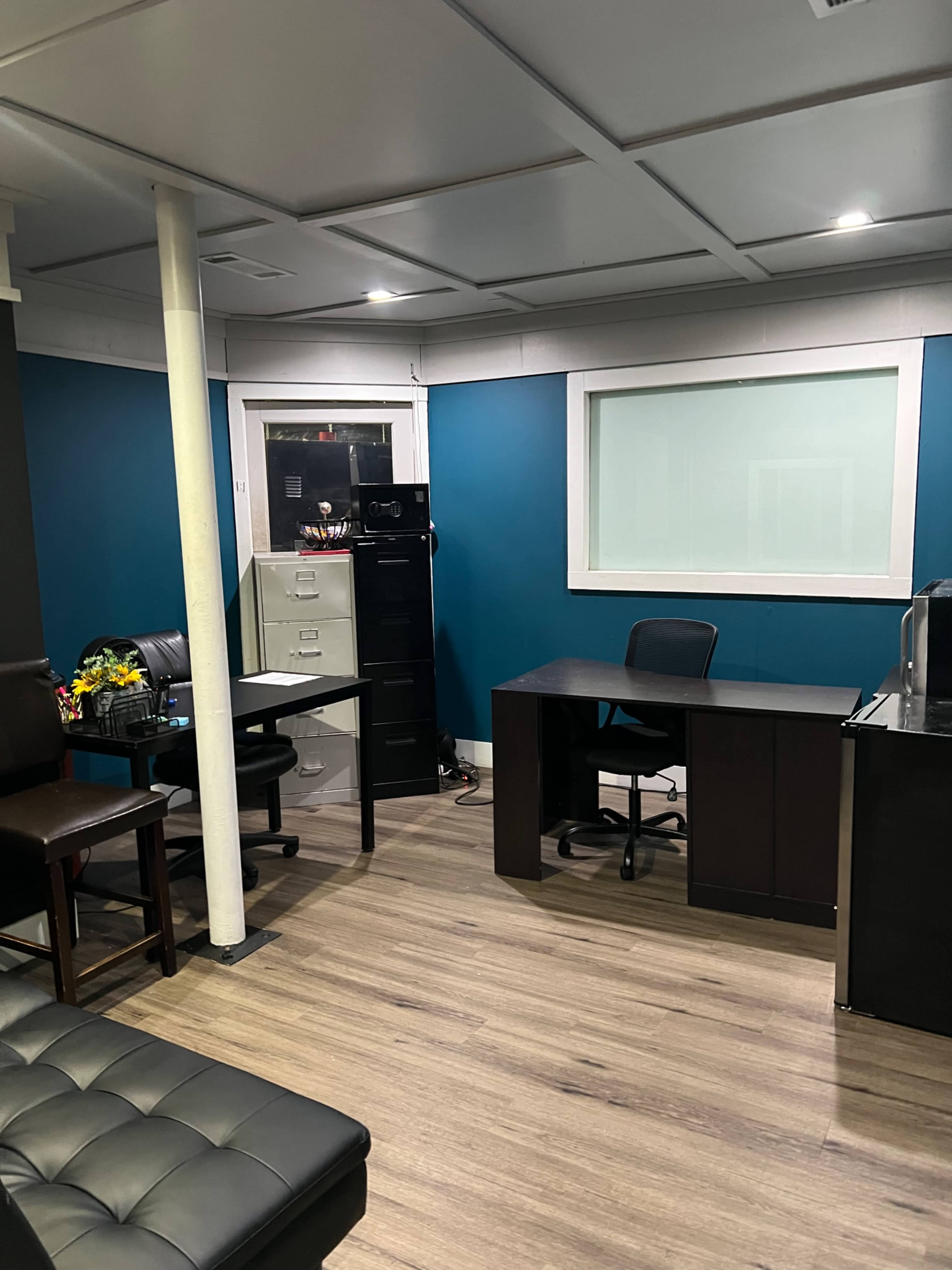 The image shows a small office space featuring a desk with a chair, a filing cabinet, and a seating area with brown chairs.