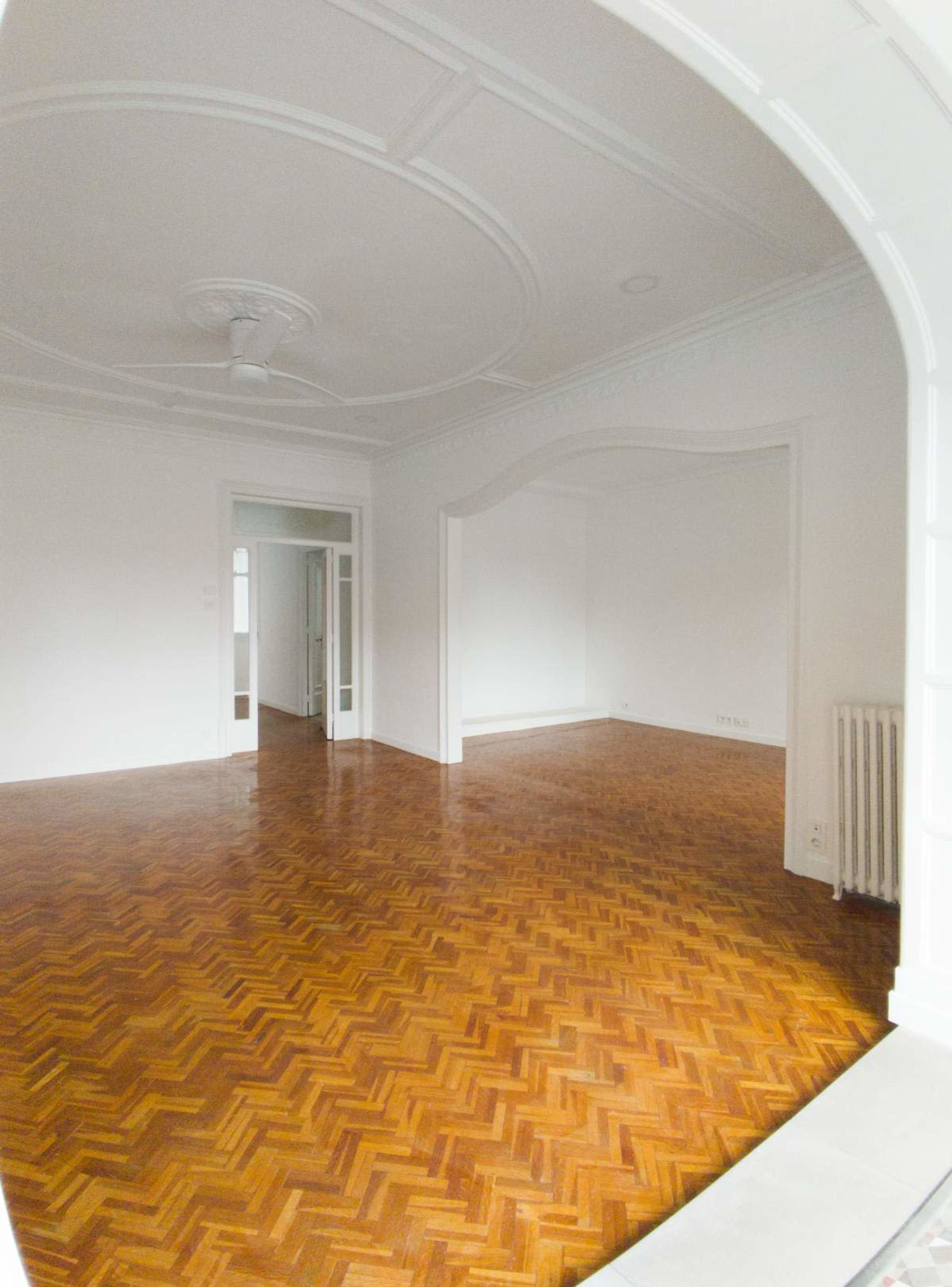 The image shows a spacious, empty room with polished herringbone wooden flooring and arched architectural details, leading to adjacent empty areas.