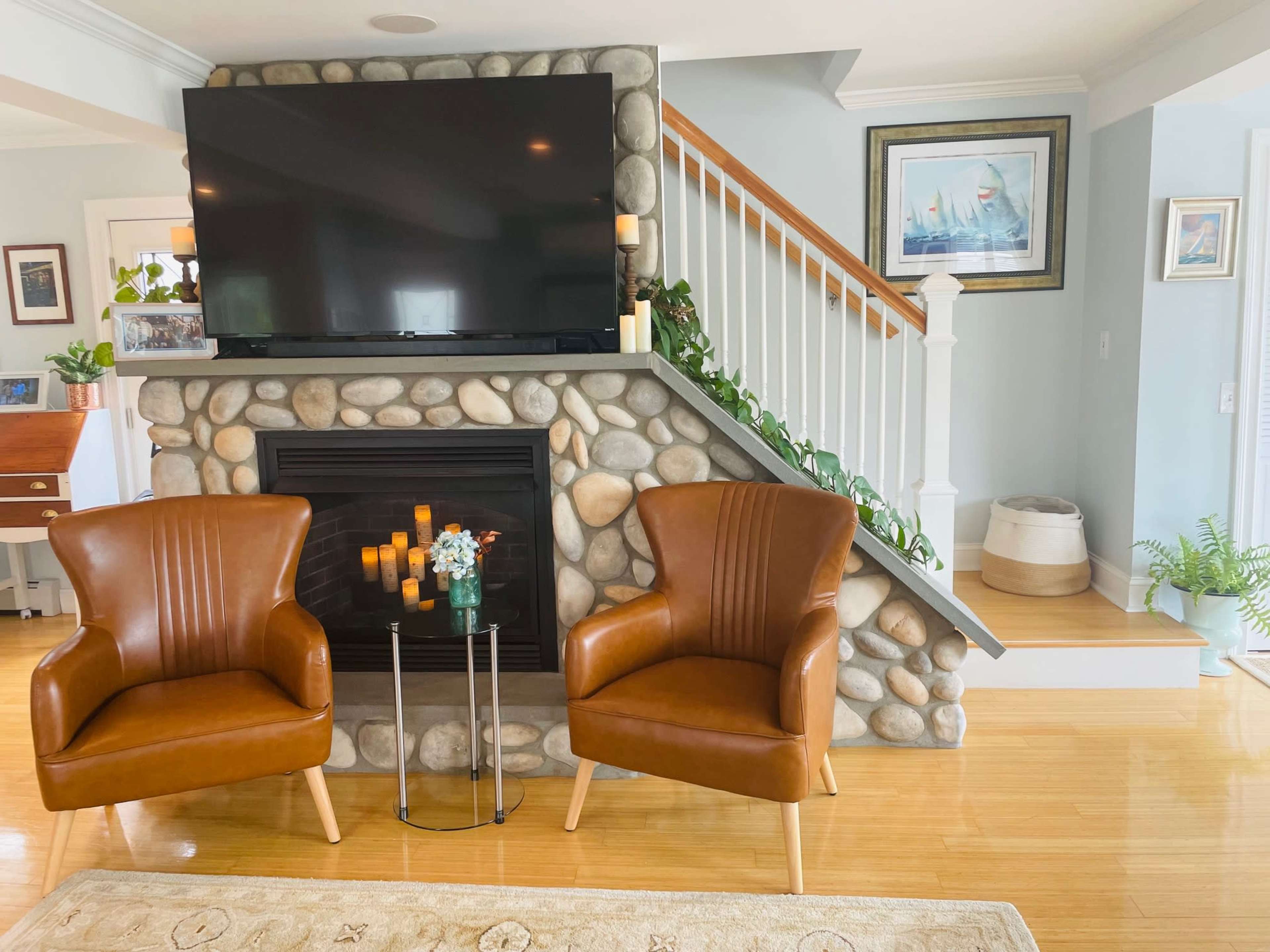 A modern living room features a stone fireplace with a large flat-screen TV above it and two brown leather chairs placed in front.
