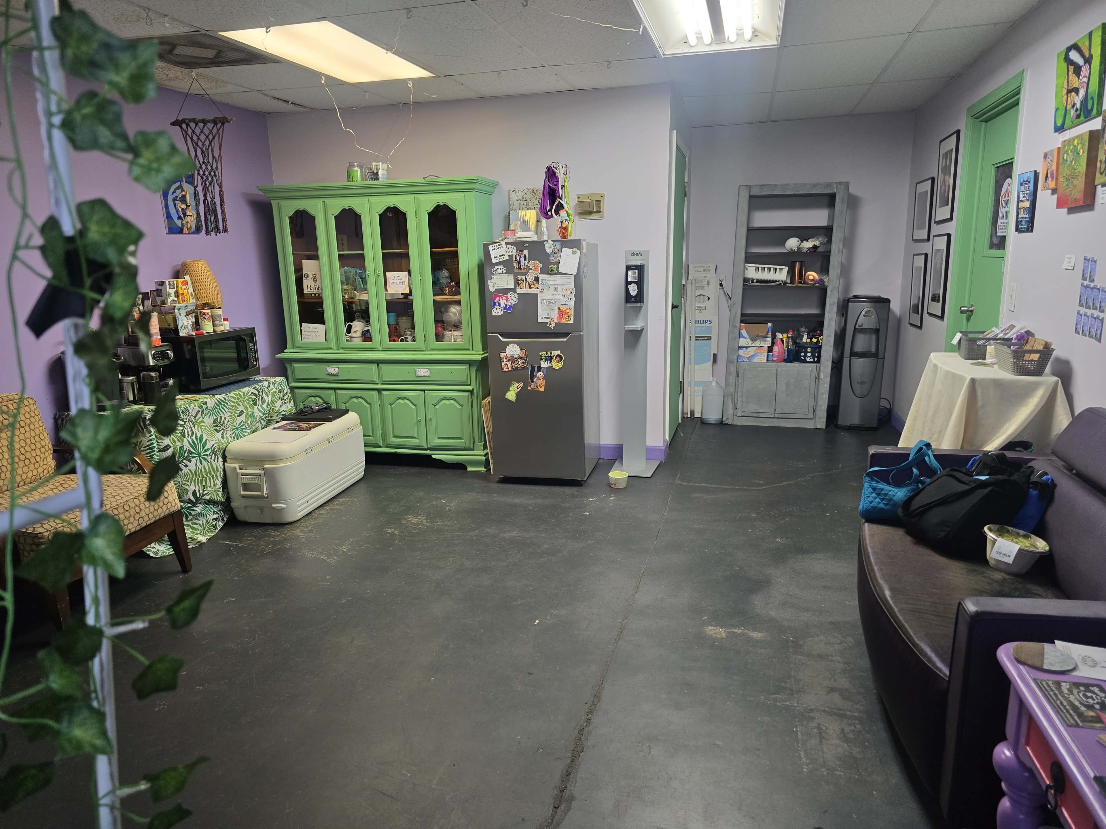 A brightly colored break room with a green cabinet, a gray refrigerator covered in magnets and notes, and a seating area featuring a couch and an armchair.