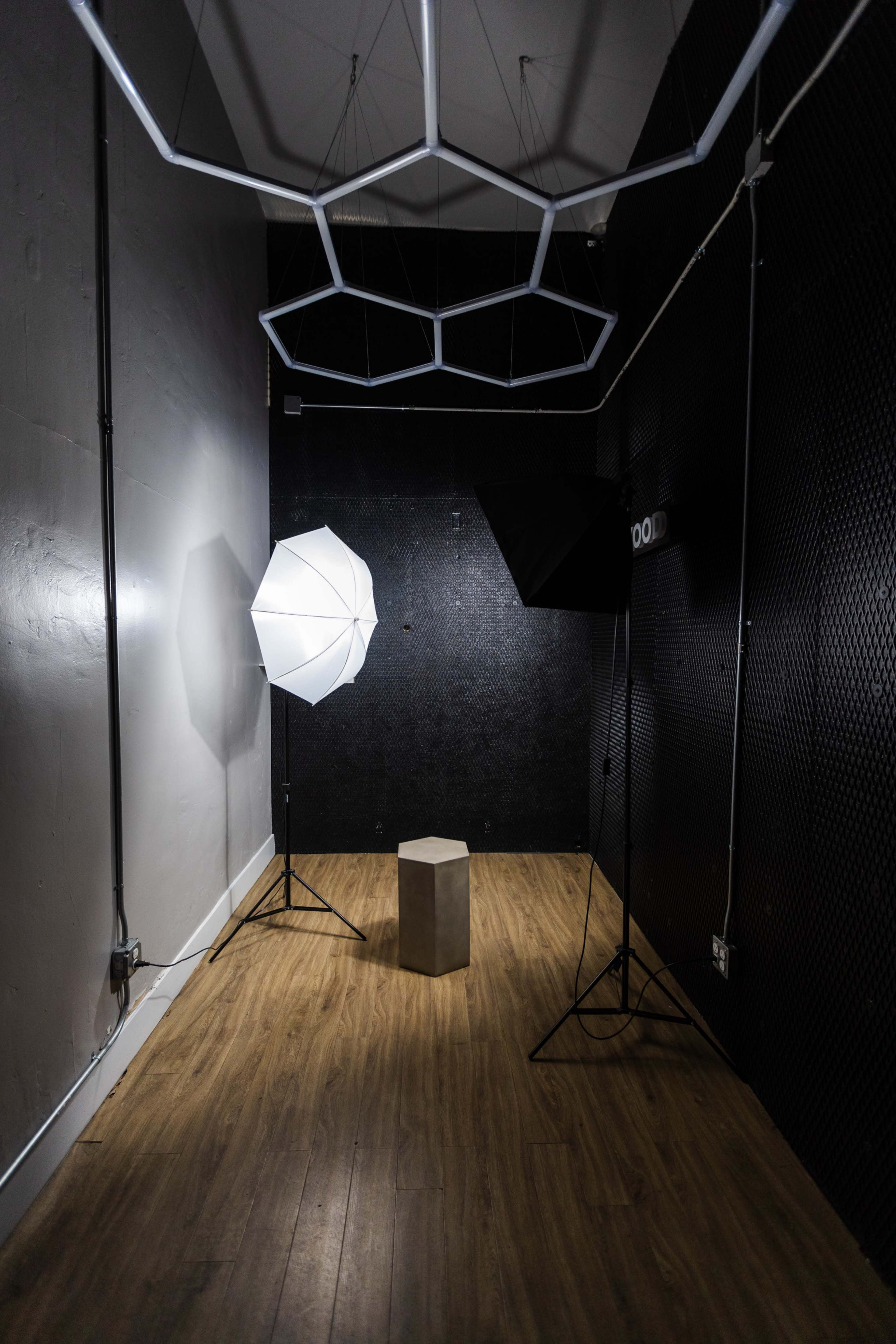 The image shows a small, empty studio space with a wooden floor, featuring two photography light setups and a gray pedestal in the center.