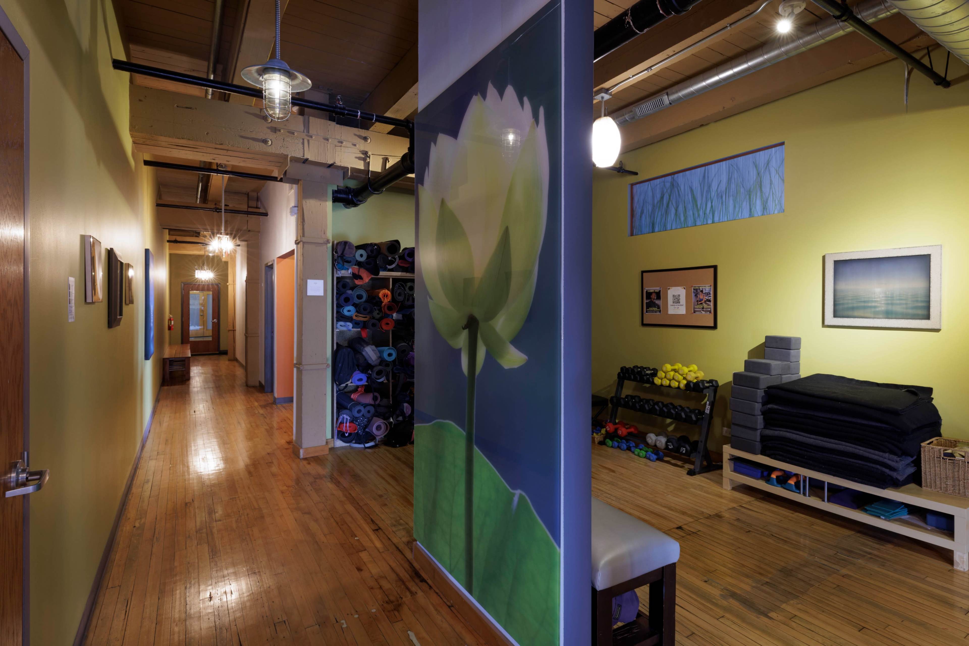 The image shows a long, narrow hallway in a fitness studio, featuring a large mural of a lotus flower and shelves stacked with yoga mats and fitness equipment along the walls.