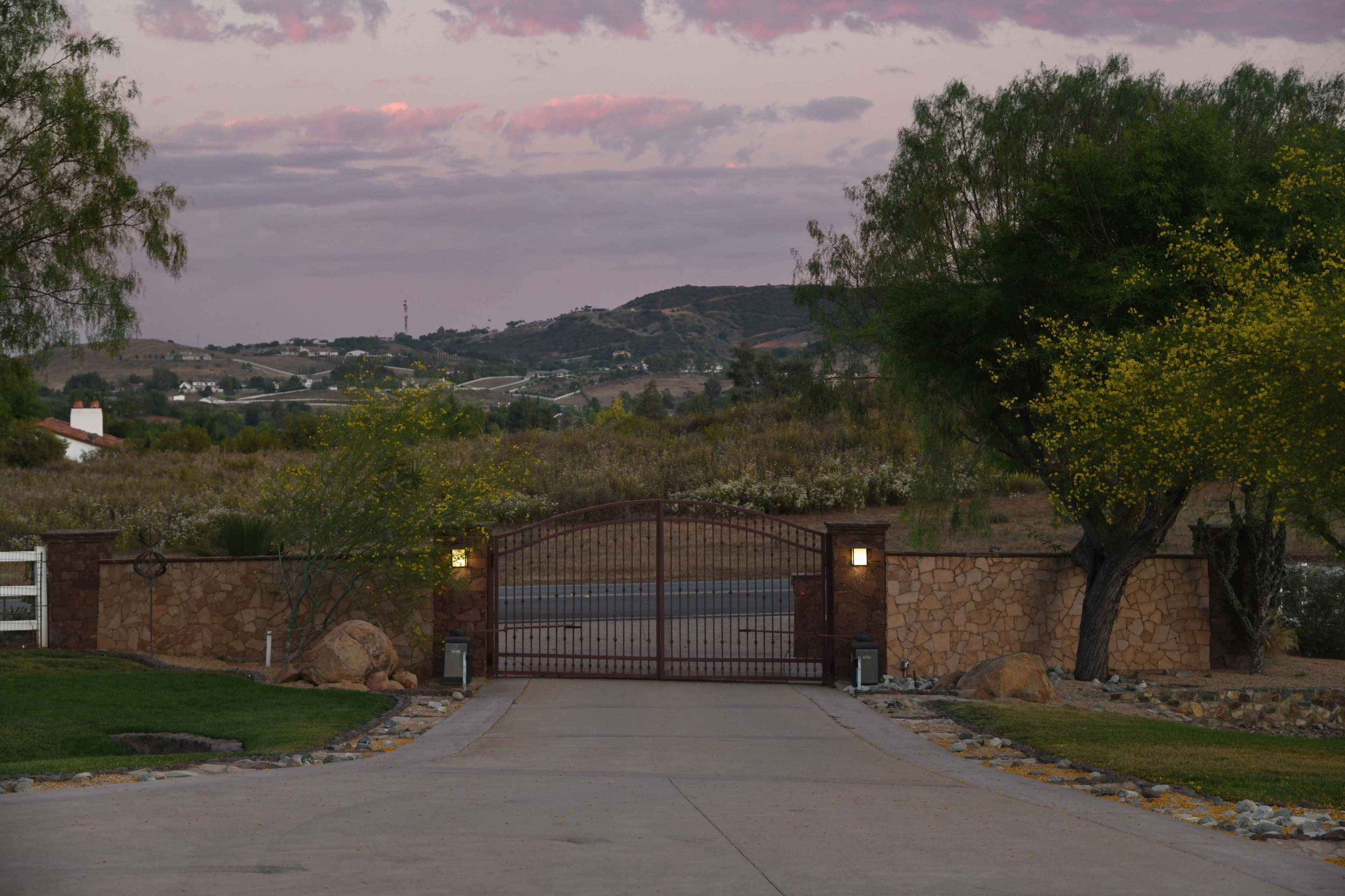 A gated entrance leads to a roadway, framed by trees and hills under a colorful sky.