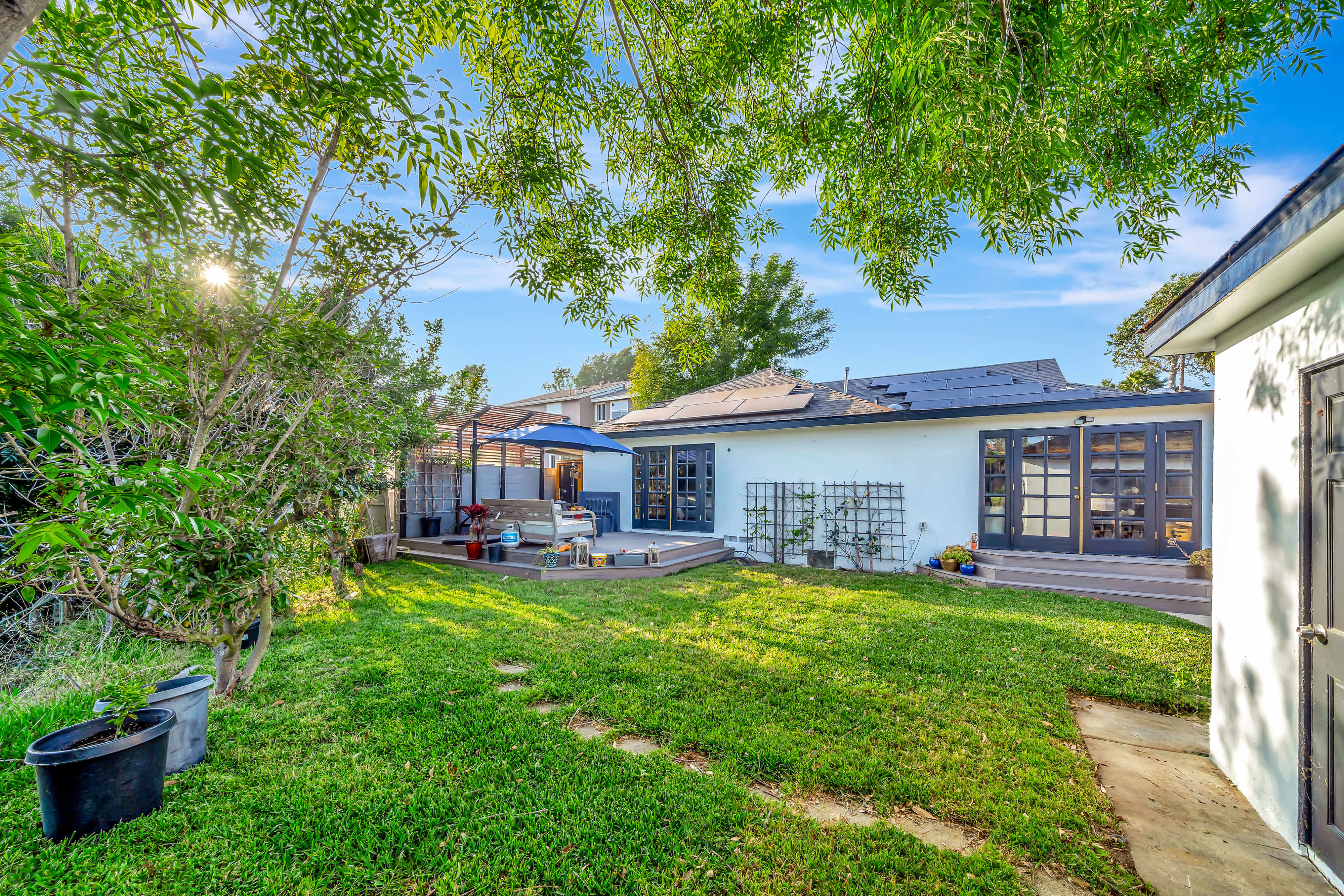The image depicts a backyard with green grass, a wooden deck, potted plants, and a seating area adjacent to a house.