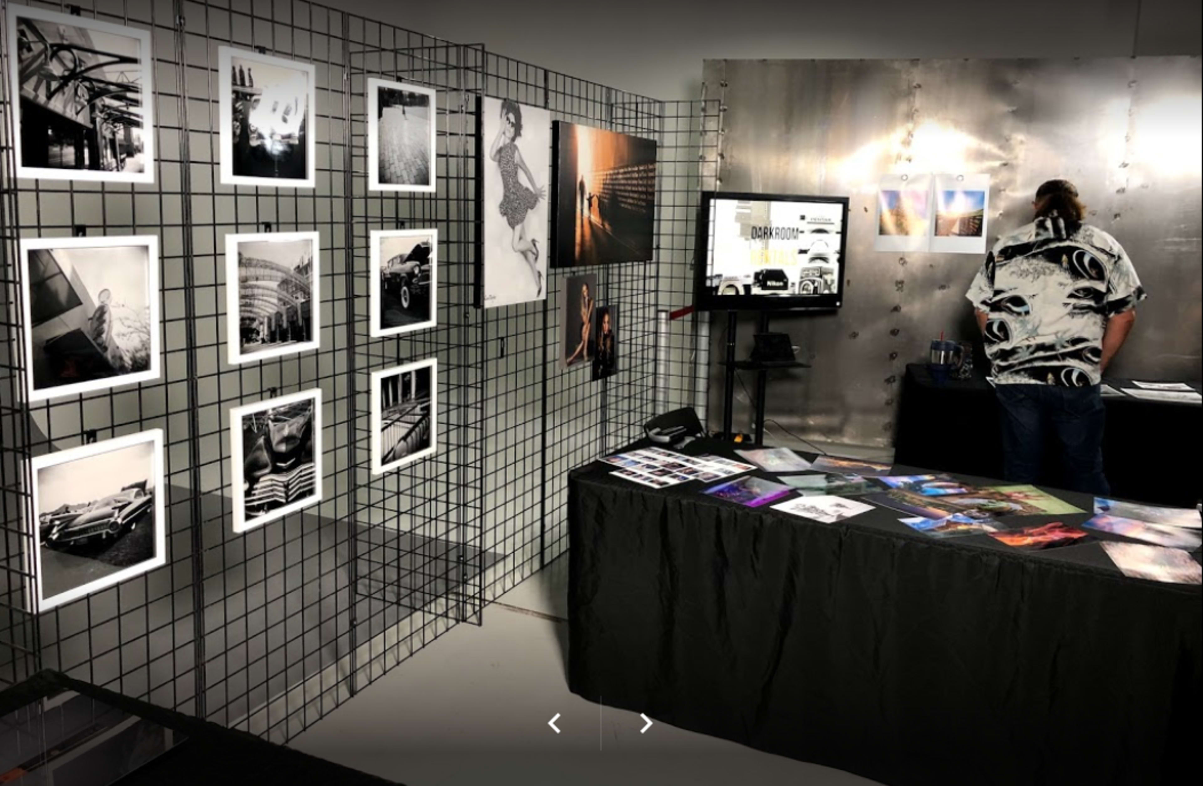A gallery display with black-and-white photographs on a grid wall and a table covered with various printed materials, while a person stands behind the table looking at a screen.