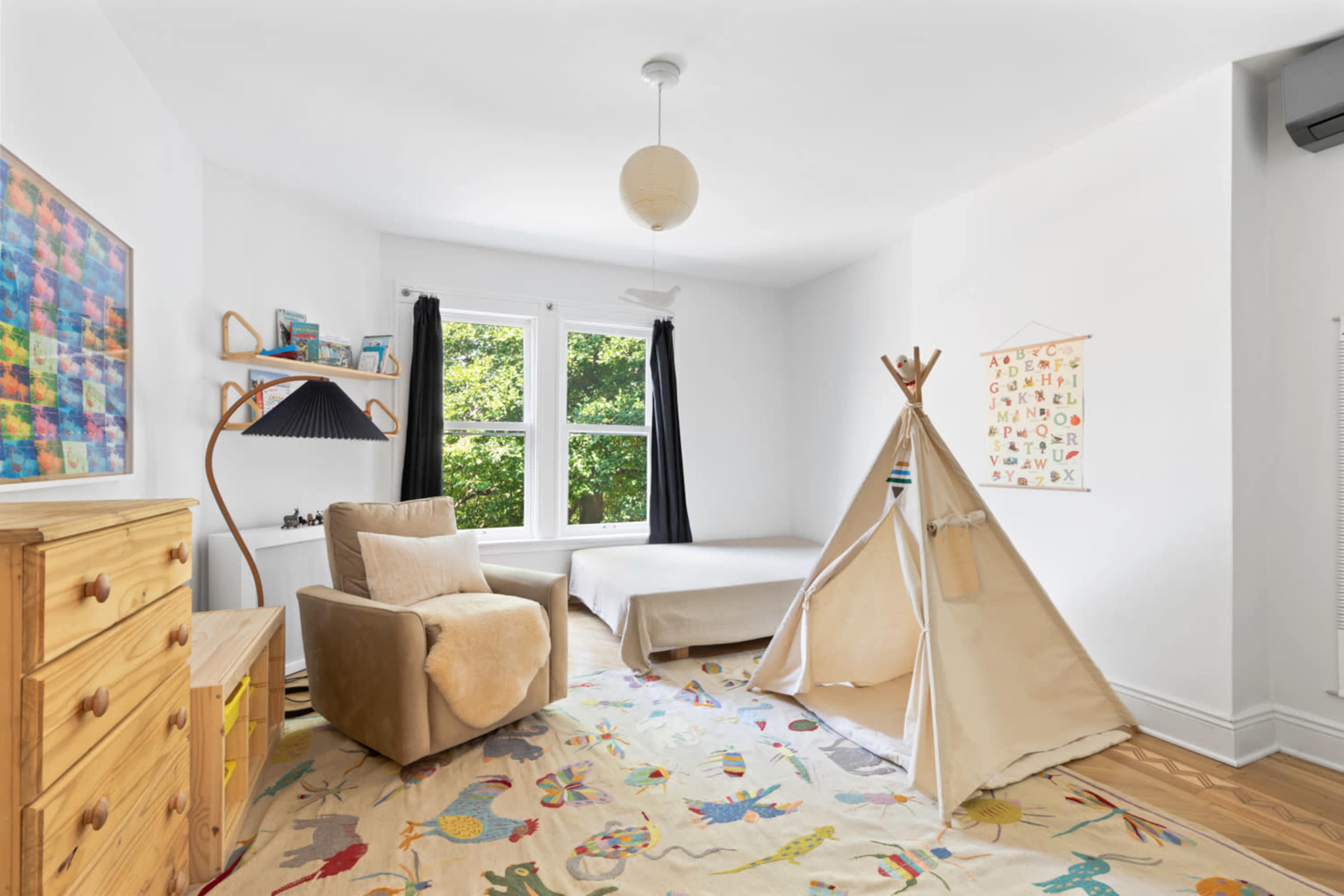The room features a single bed, a cozy armchair, a decorative teepee, and a colorful area rug, with large windows allowing natural light to enter.
