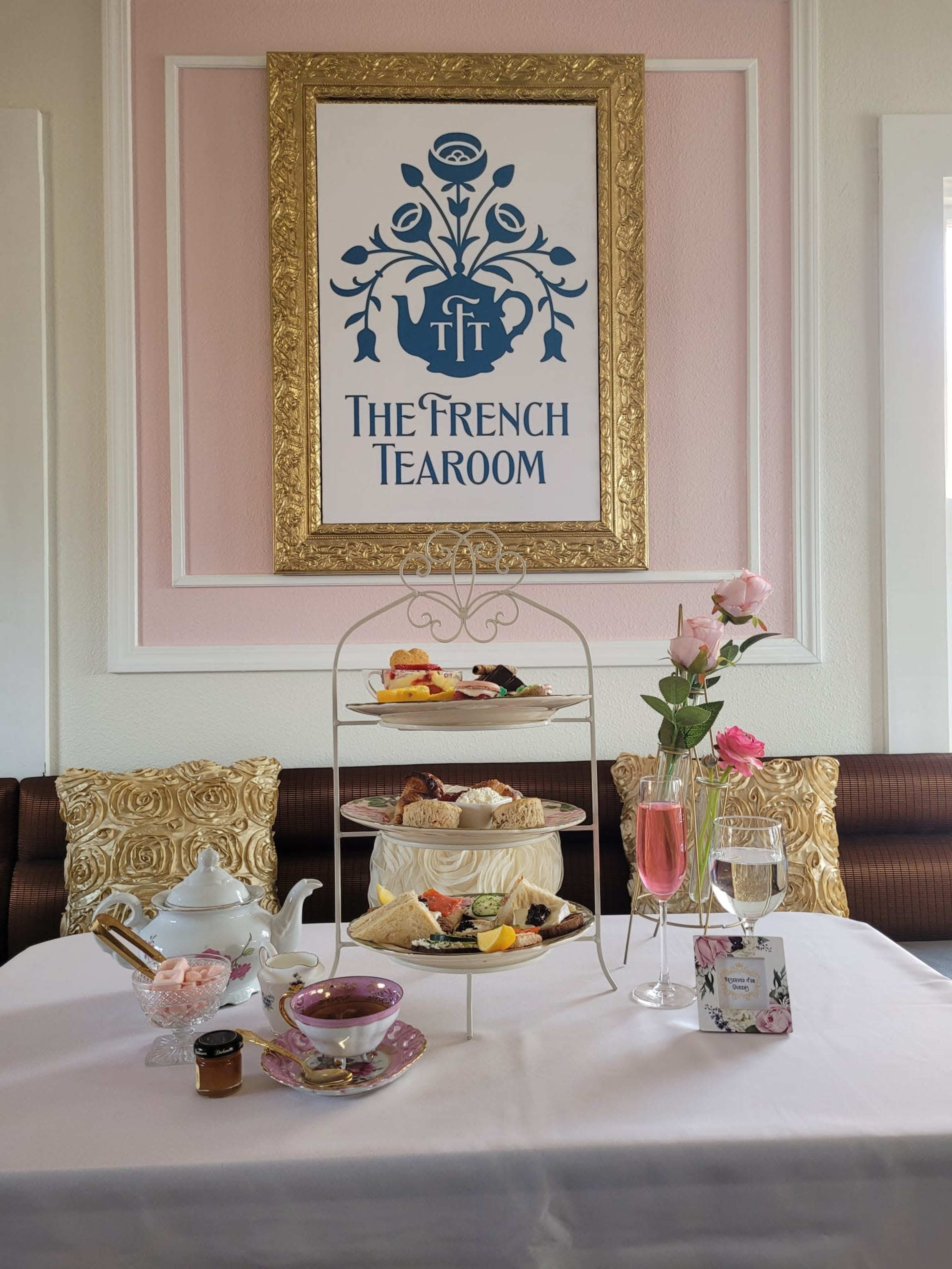 A table is elegantly set with a tiered stand of assorted pastries and sandwiches, accompanied by a teapot, glasses of sparkling water, and fresh roses, all under a decorative sign that reads "The French Tearoom."