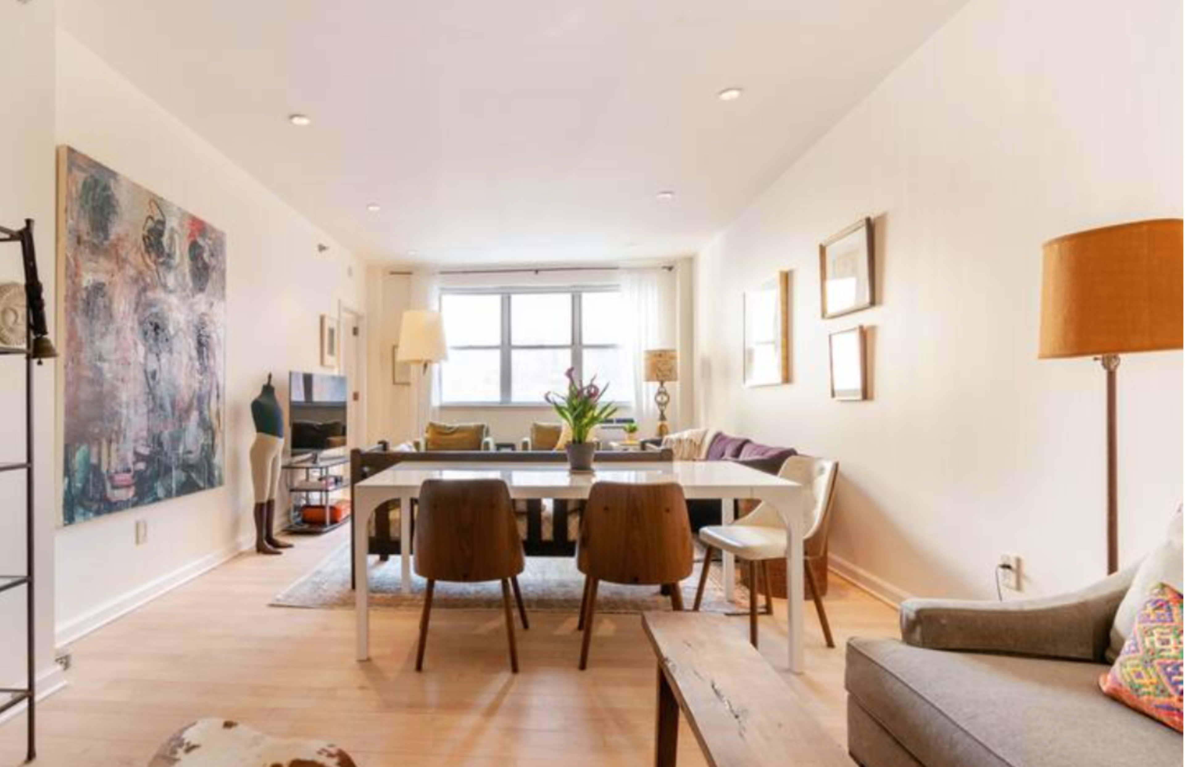 A modern living room and dining area featuring a large painting, a dining table with chairs, and a seating arrangement with a couch and lamps.