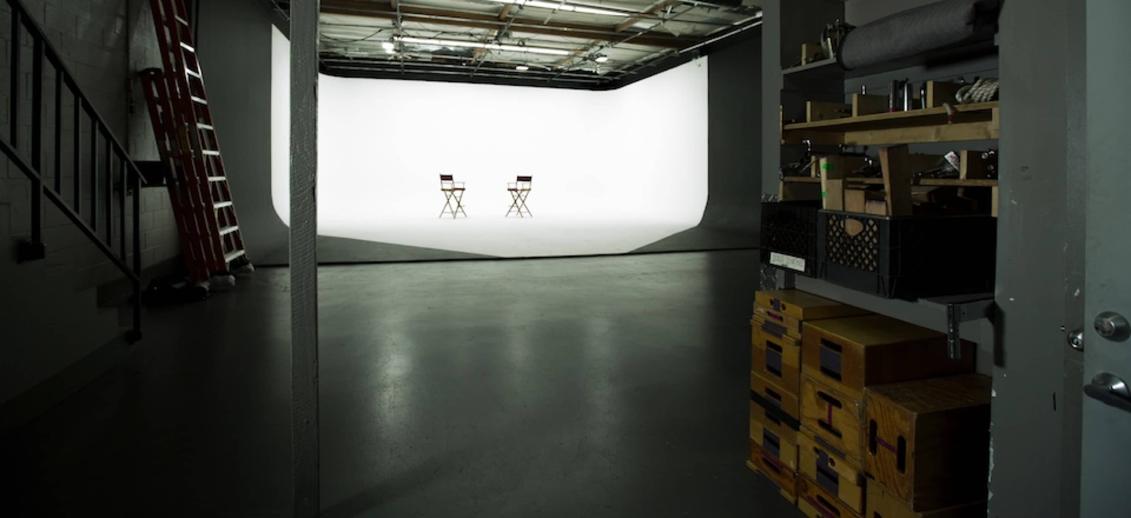 An empty studio space with two director's chairs facing a white backdrop, flanked by shelves containing equipment.