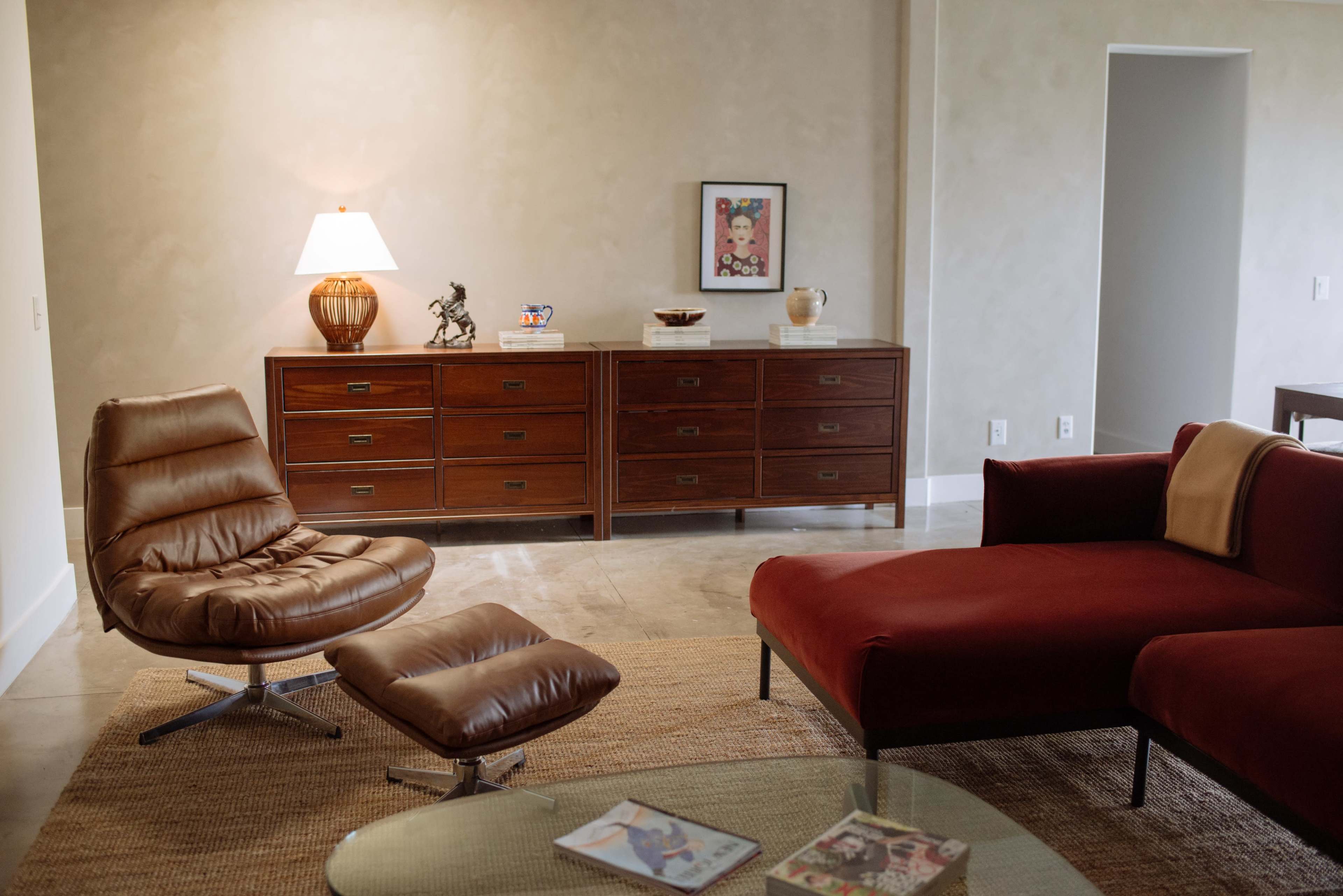The image depicts a modern living room with a leather chair and ottoman, a red sectional sofa, a wooden dresser, and a lamp on a side table, set against a neutral-colored wall.