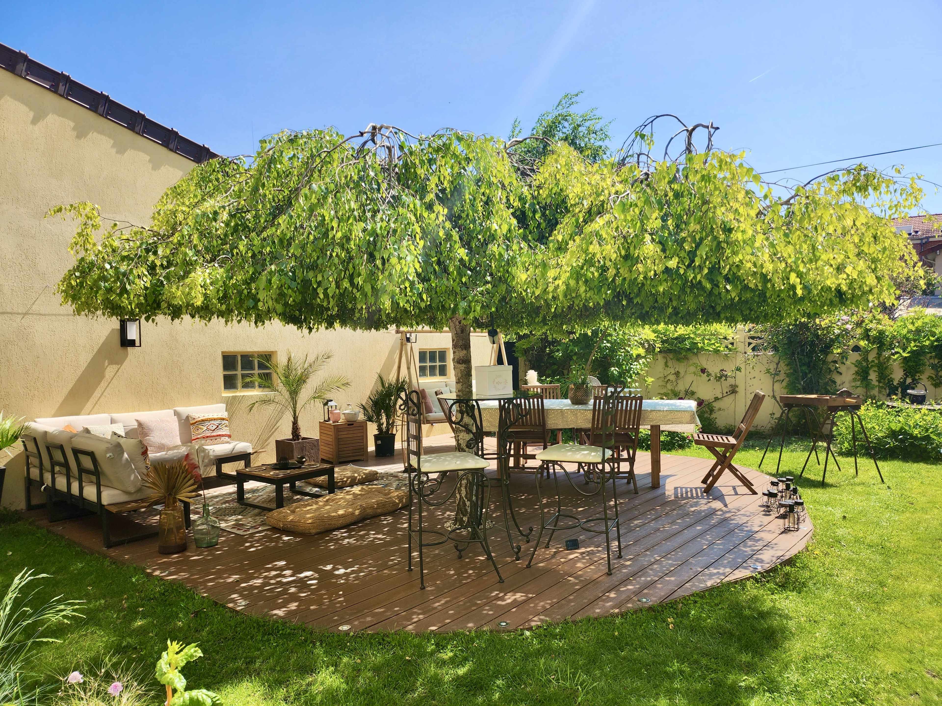 A wooden deck features outdoor furniture under a wide canopy of greenery in a well-maintained backyard.