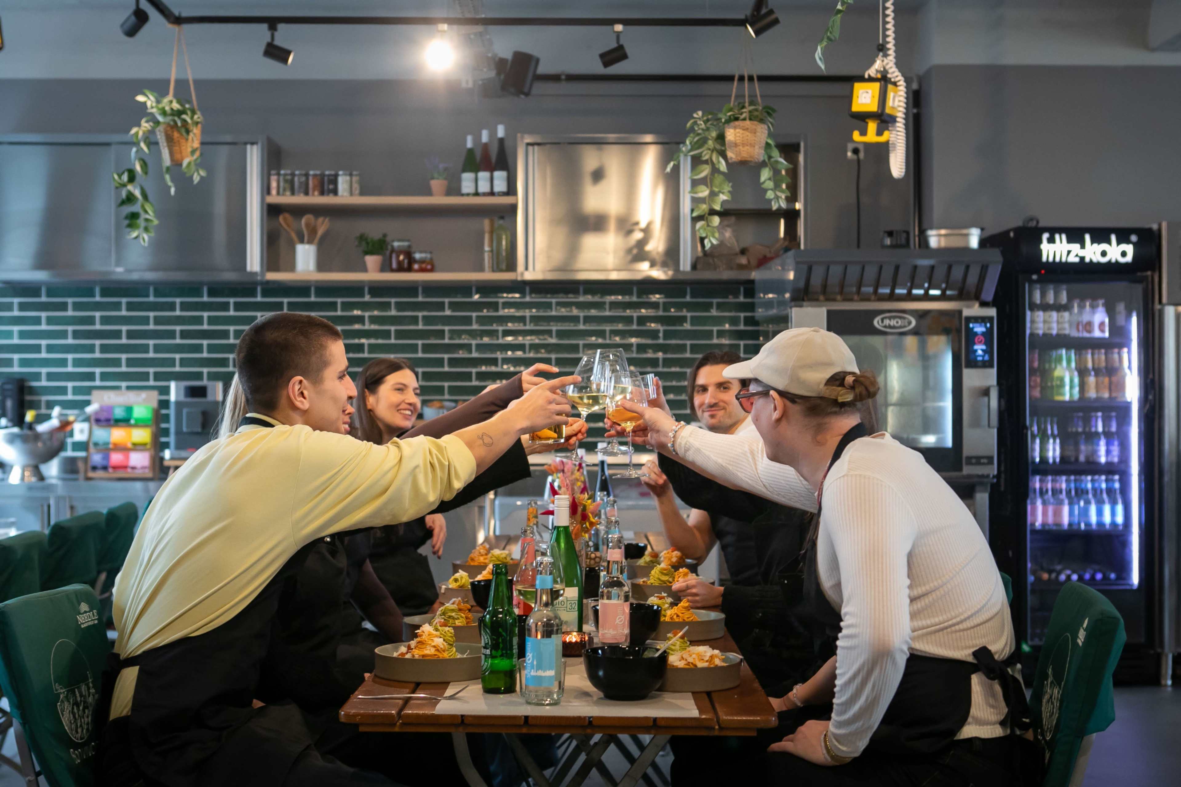 A group of five people are seated at a long table, raising their glasses in a toast amidst a restaurant setting with food and drinks arranged in front of them.