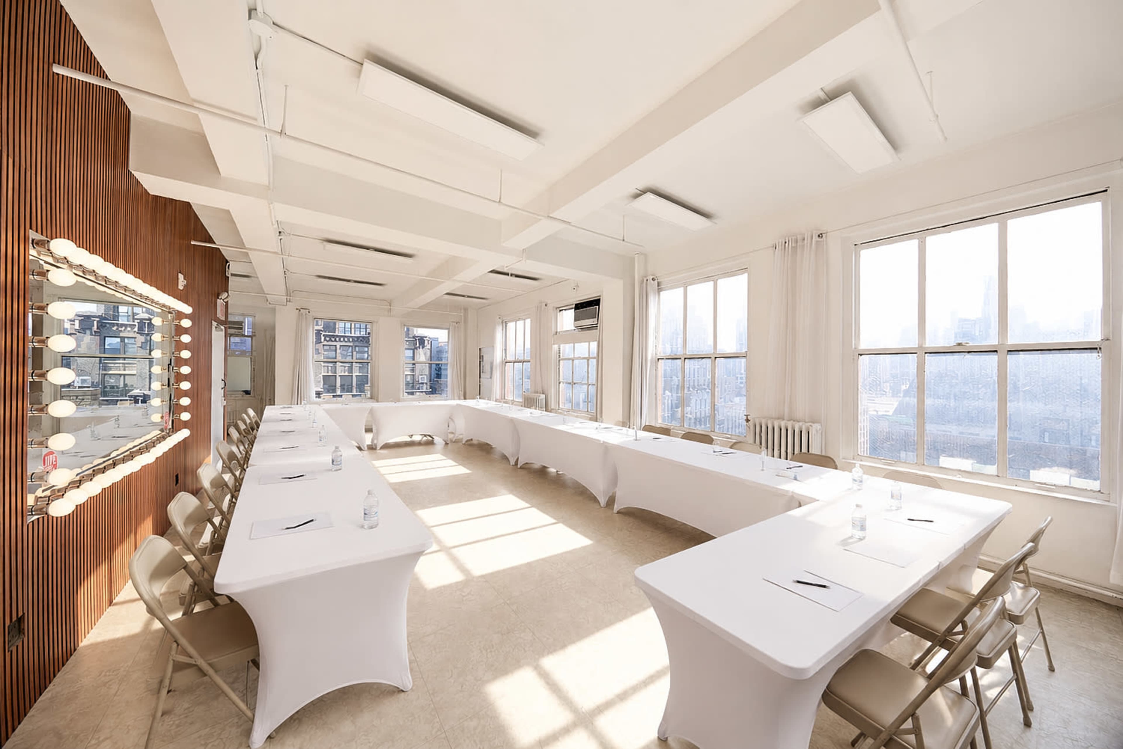 The image shows a bright, empty conference room with two long tables arranged in a U-shape and large windows allowing natural light to fill the space.