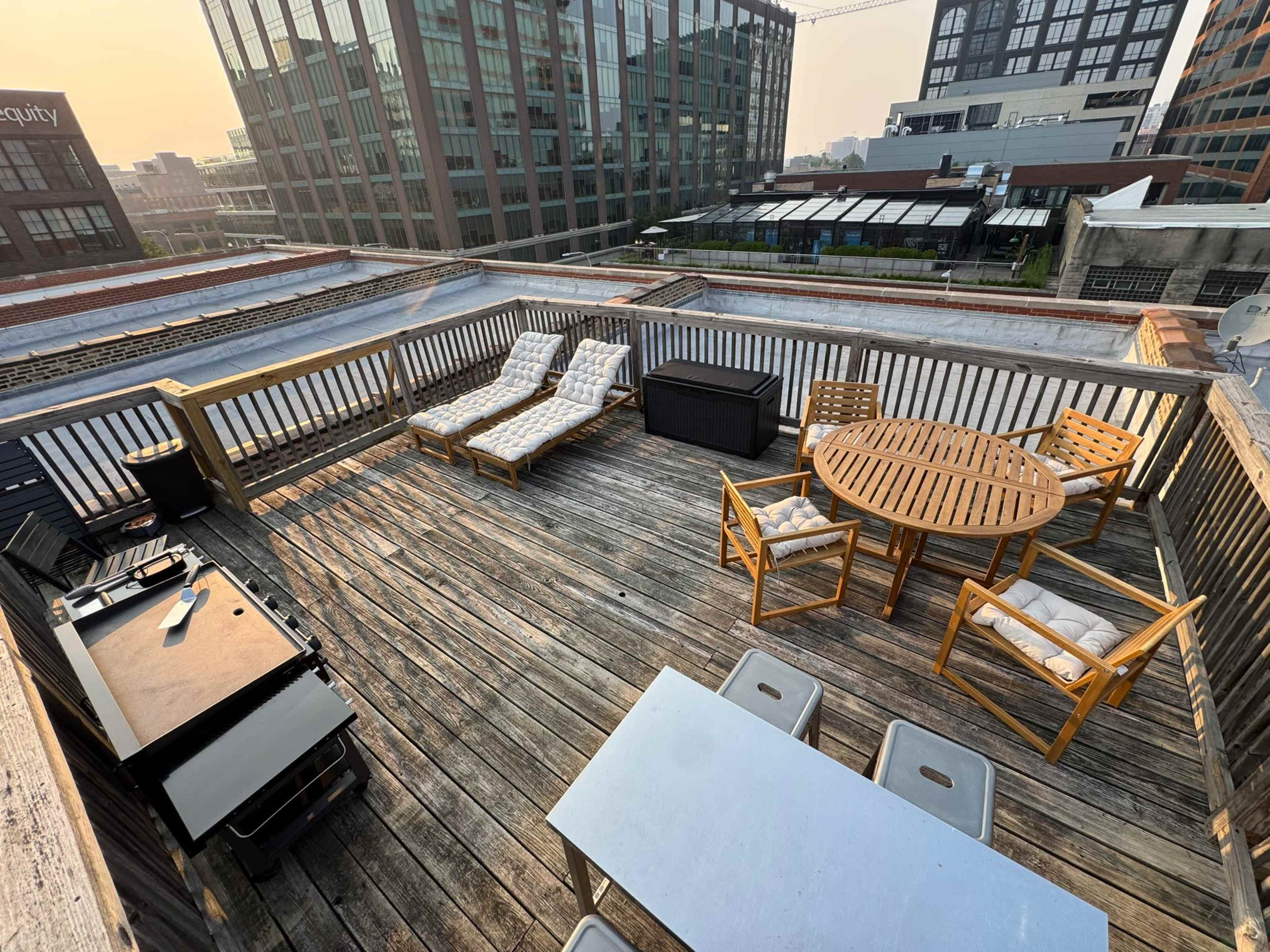 The image shows a rooftop patio featuring wooden decking, lounge chairs, a round dining table, an outdoor grill, and various urban buildings in the background.