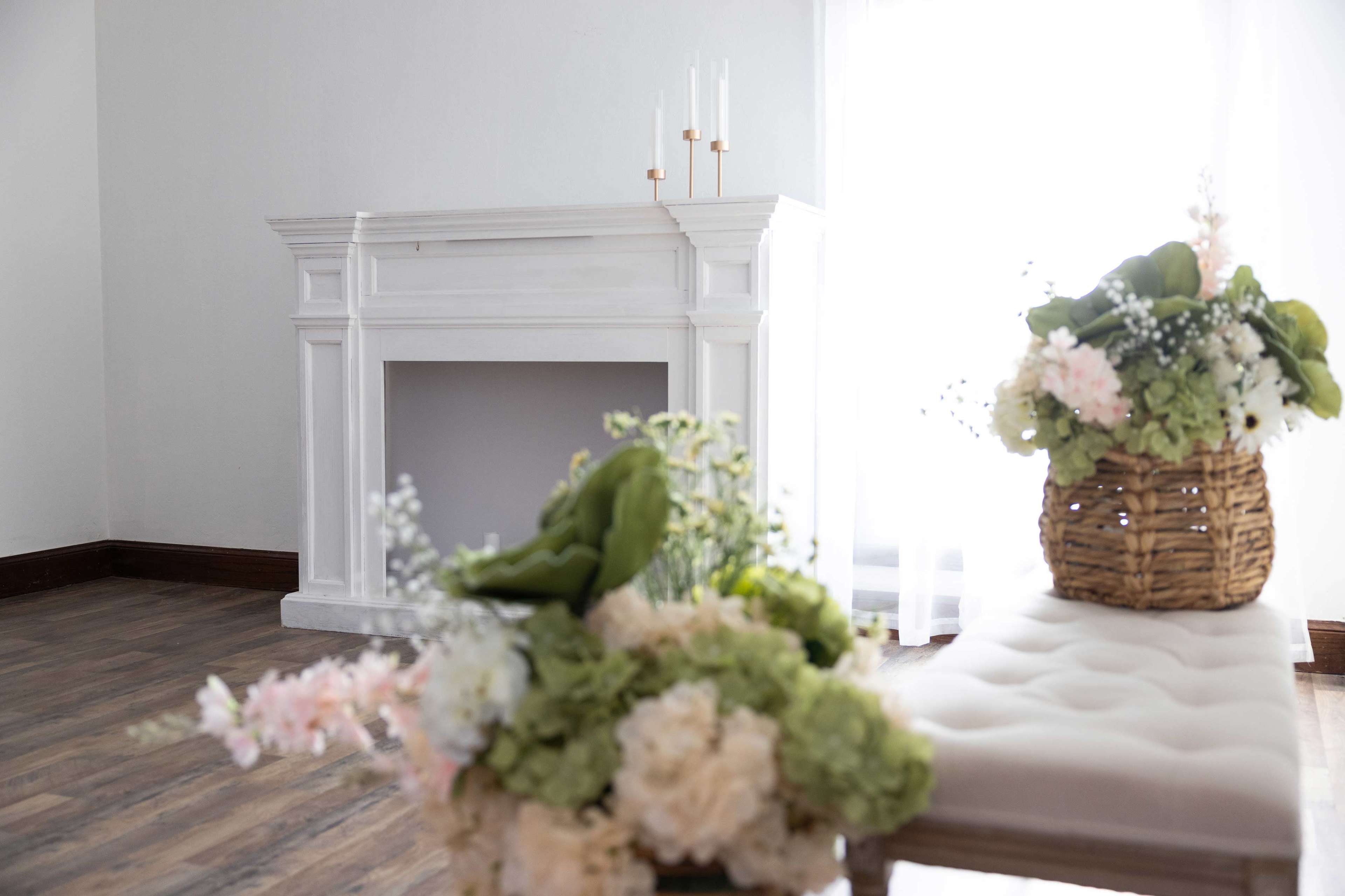 A bright room features a white mantelpiece and a bench adorned with baskets of fresh flowers.