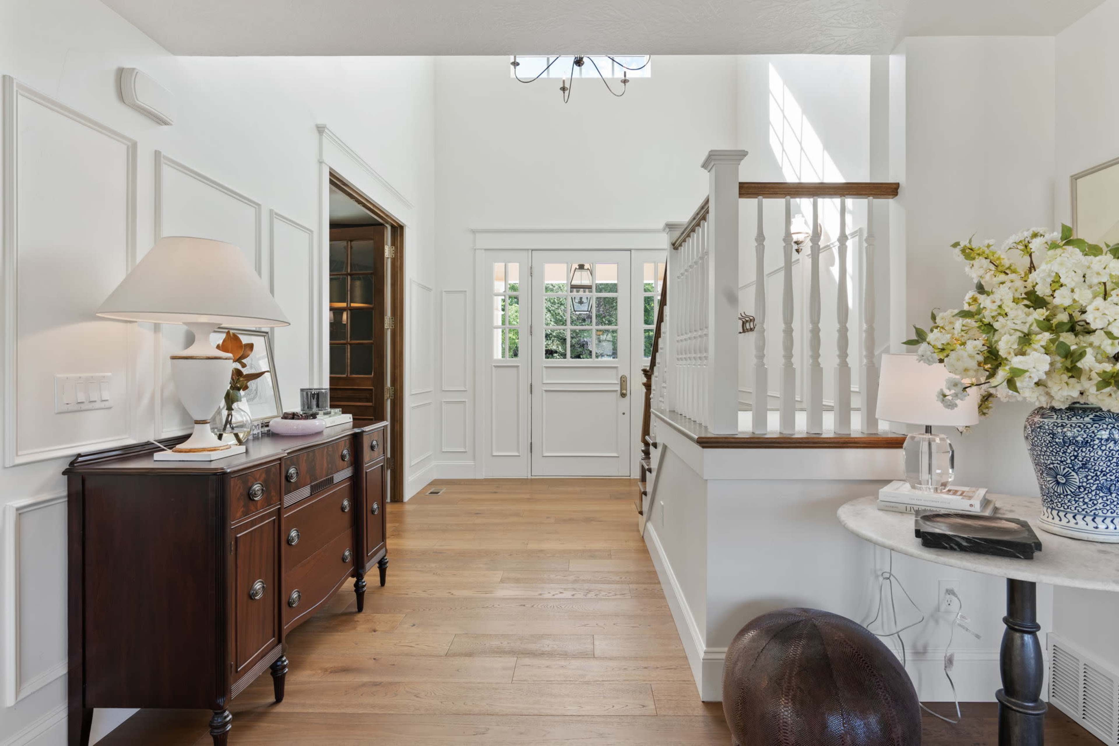 A bright entryway featuring a staircase, a wooden sideboard, and decorative elements like a lamp and a vase of flowers.