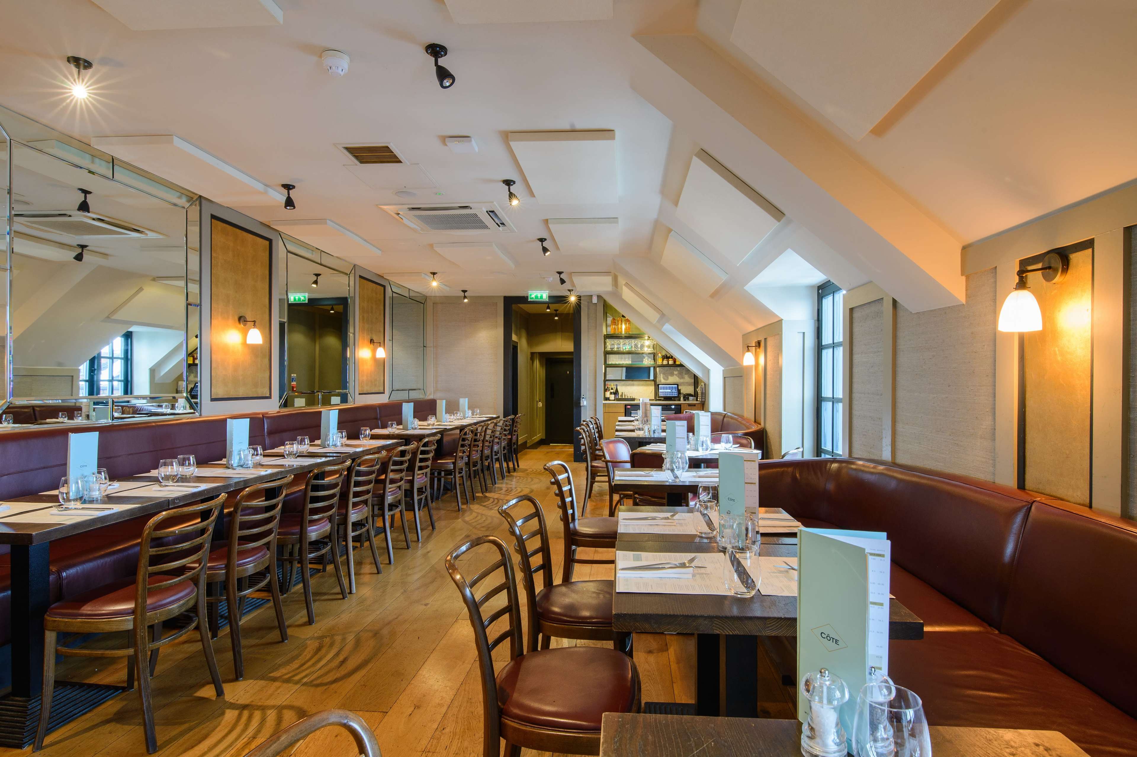 The image shows a spacious restaurant interior with wooden tables and brown leather seating arranged in rows under a sloped ceiling.