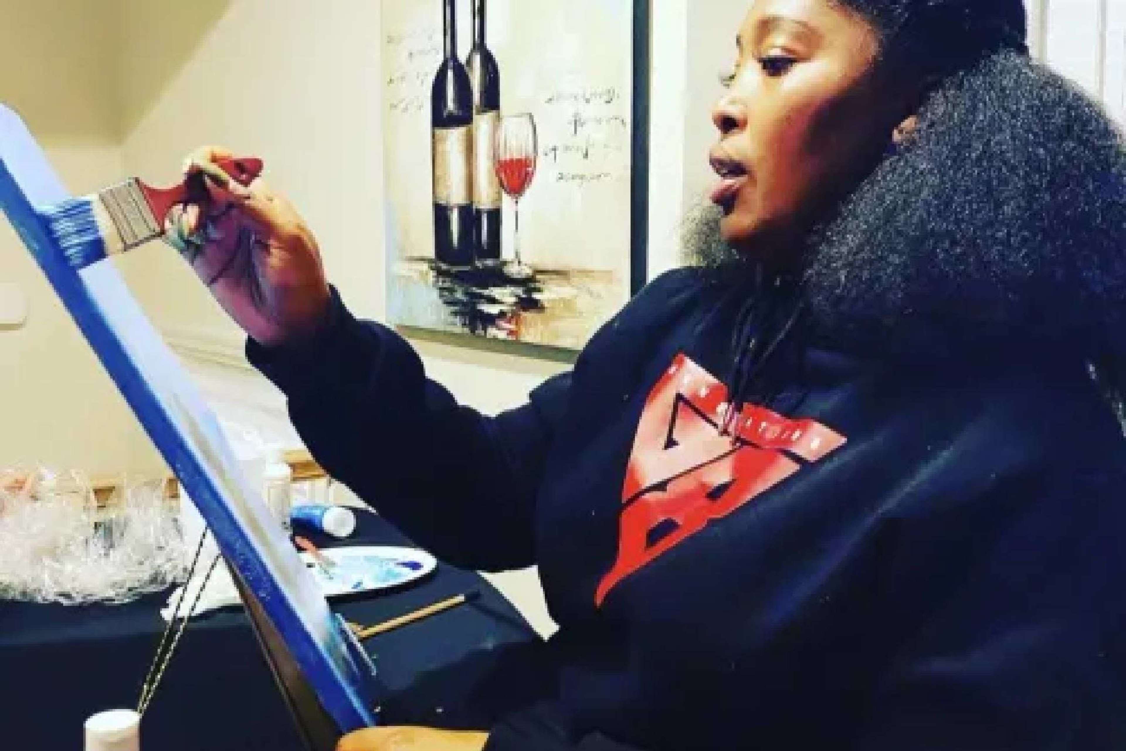 A person is painting on a canvas while seated in a room with wine-themed artwork in the background.