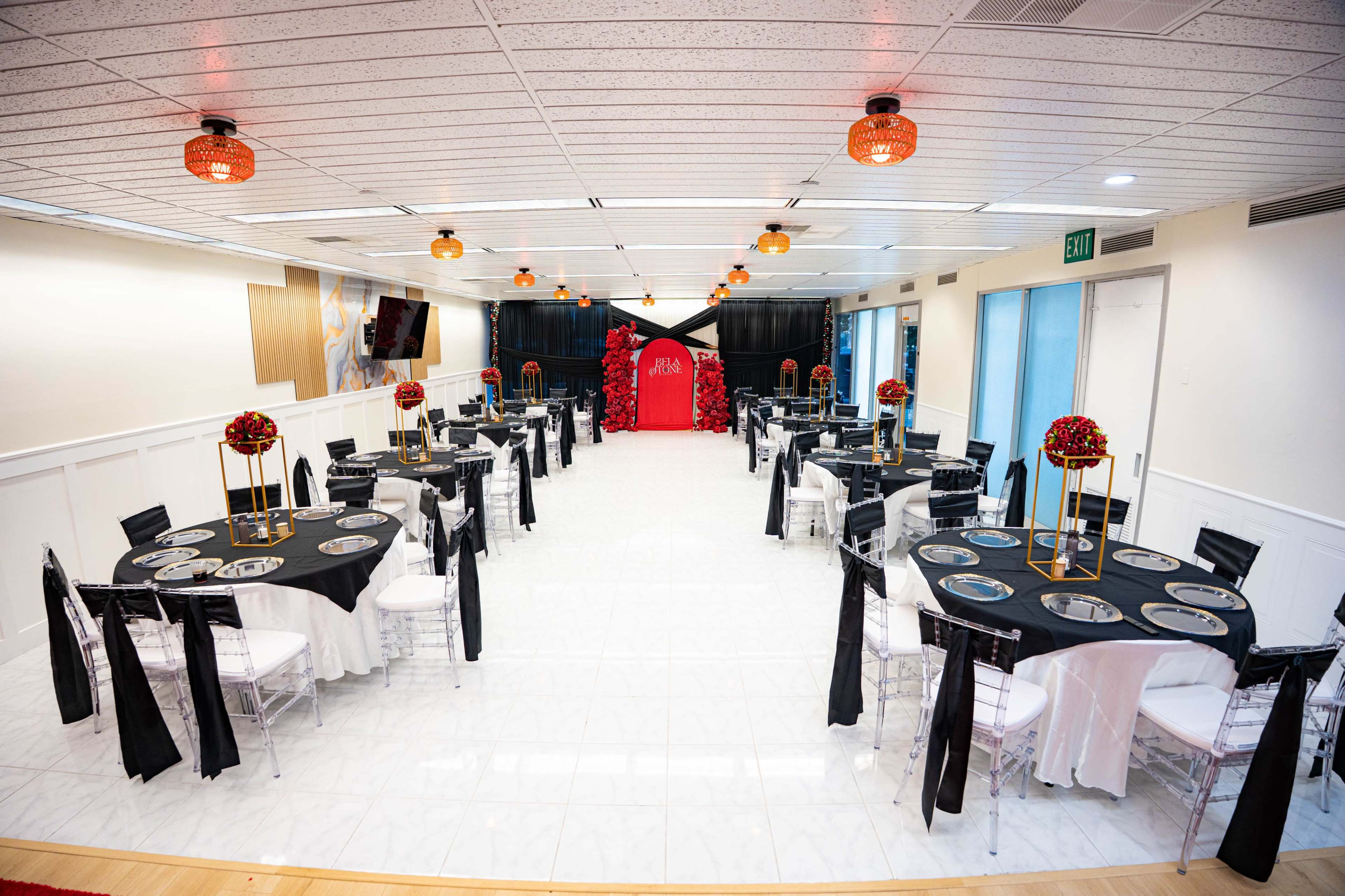 The image shows an elegantly arranged event space with round tables covered in black and white tablecloths, featuring decorative centerpieces, and a backdrop in red at the front.