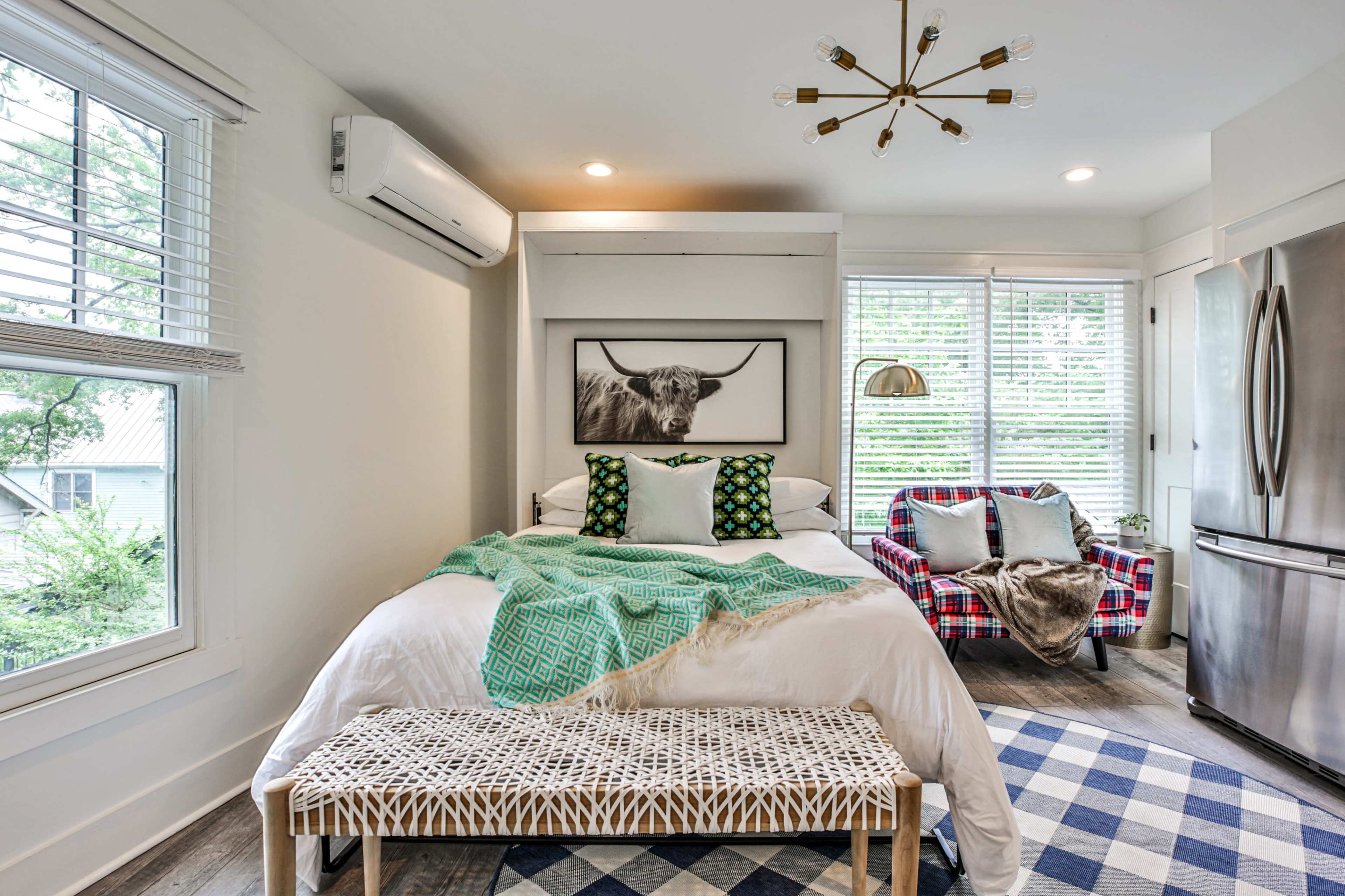 A small bedroom with a bed, a plaid chair, a window with blinds, and a refrigerator in the corner.