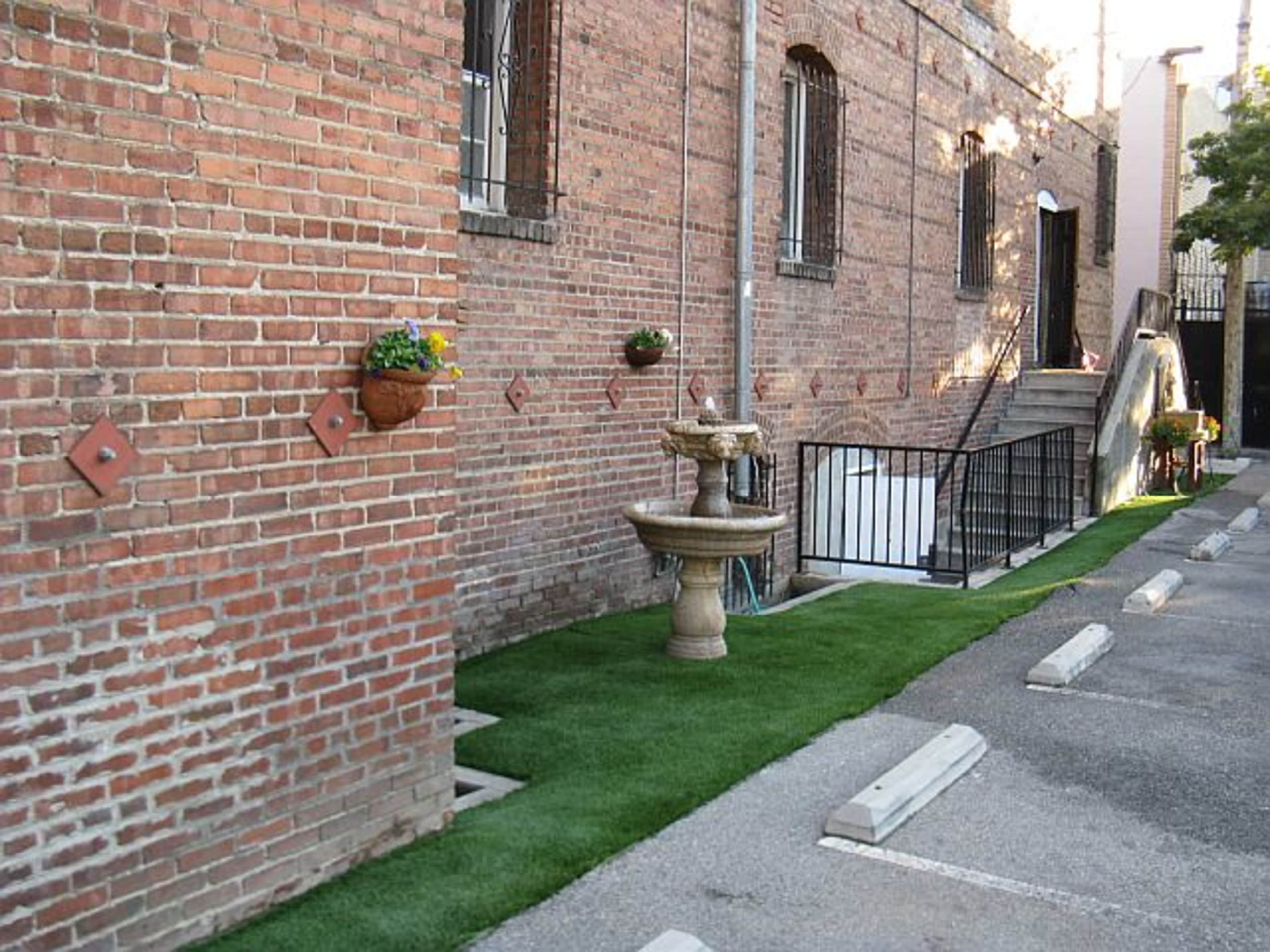 A brick wall beside a walkway featuring a fountain and potted plants, alongside a strip of artificial turf and parking spaces.