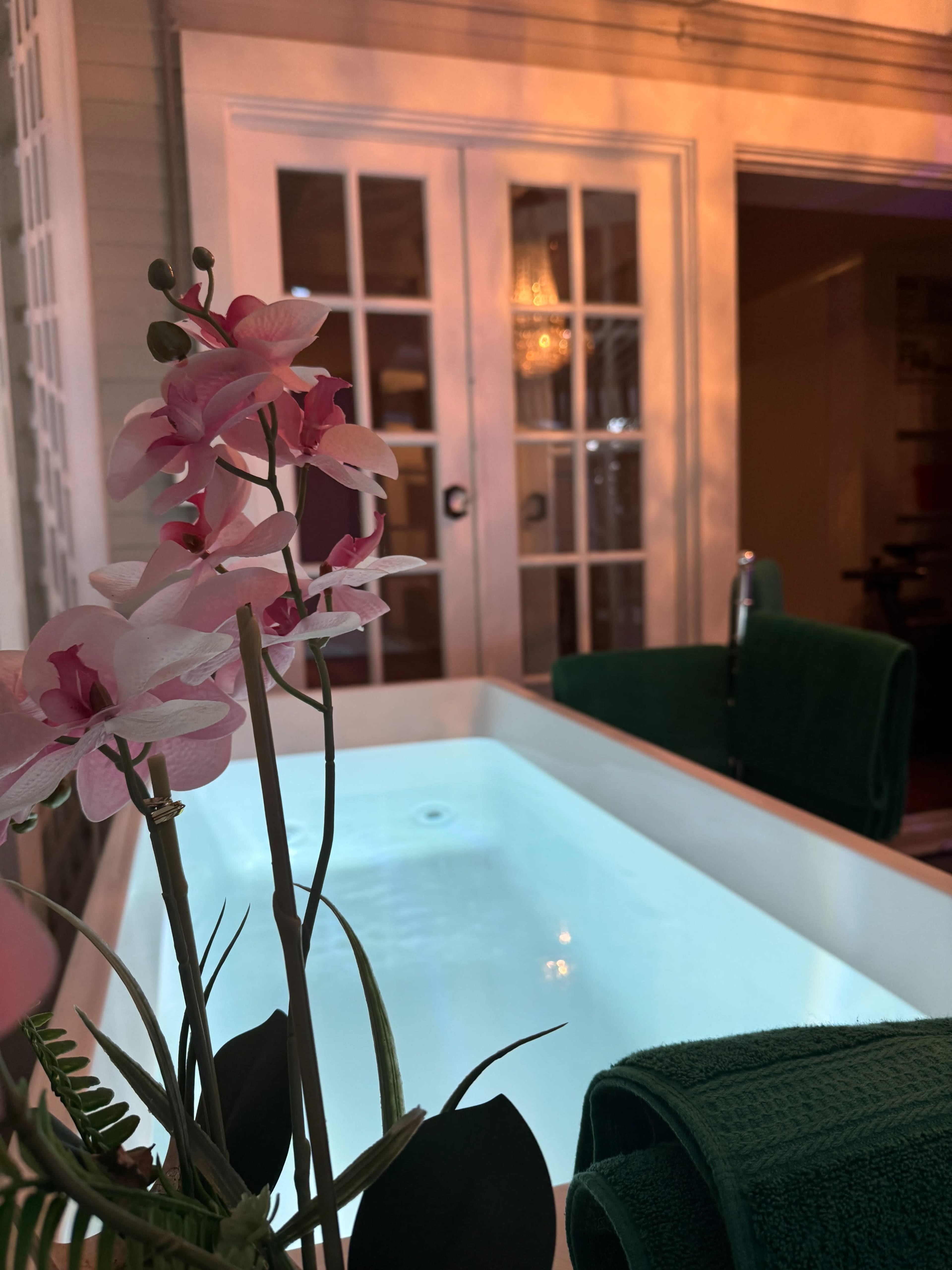 A white spa tub with softly lit water is positioned near a flowering plant and green towels, set against a backdrop of glass doors.