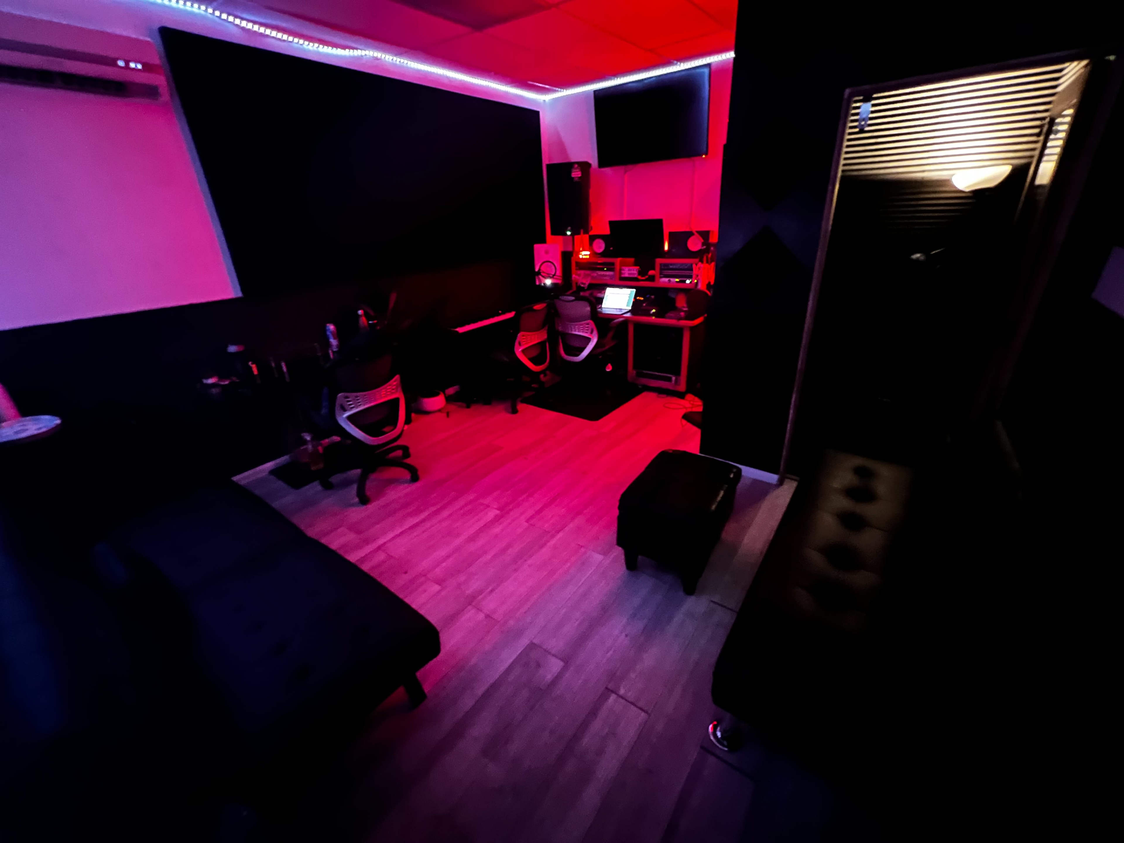 A dimly lit recording studio with black walls, two office chairs, a computer setup, and a small seating area.