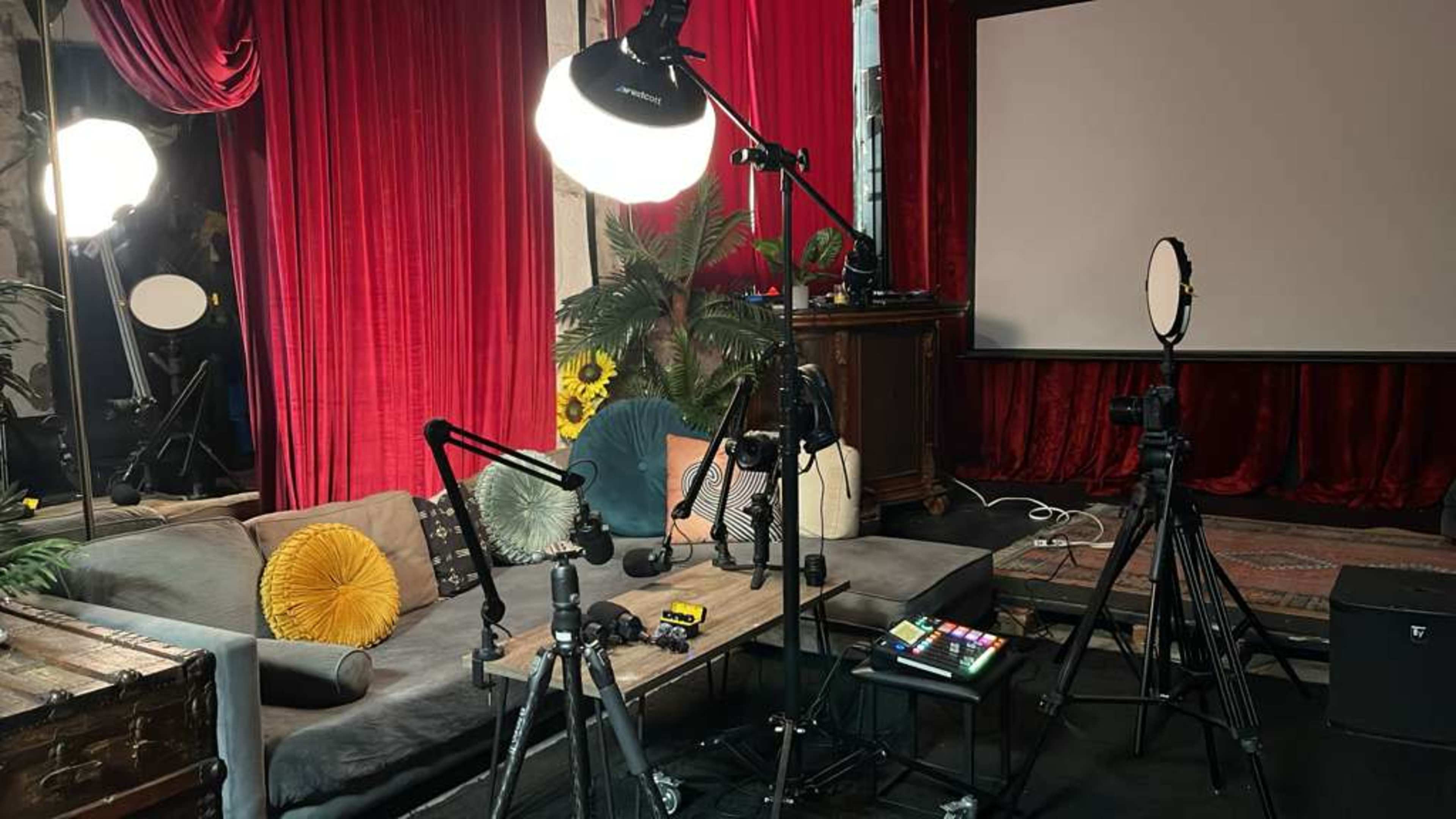 The image shows a styled indoor studio setup featuring a gray sofa with decorative cushions, various lighting equipment, a projector screen in the background, and tripods positioned nearby.