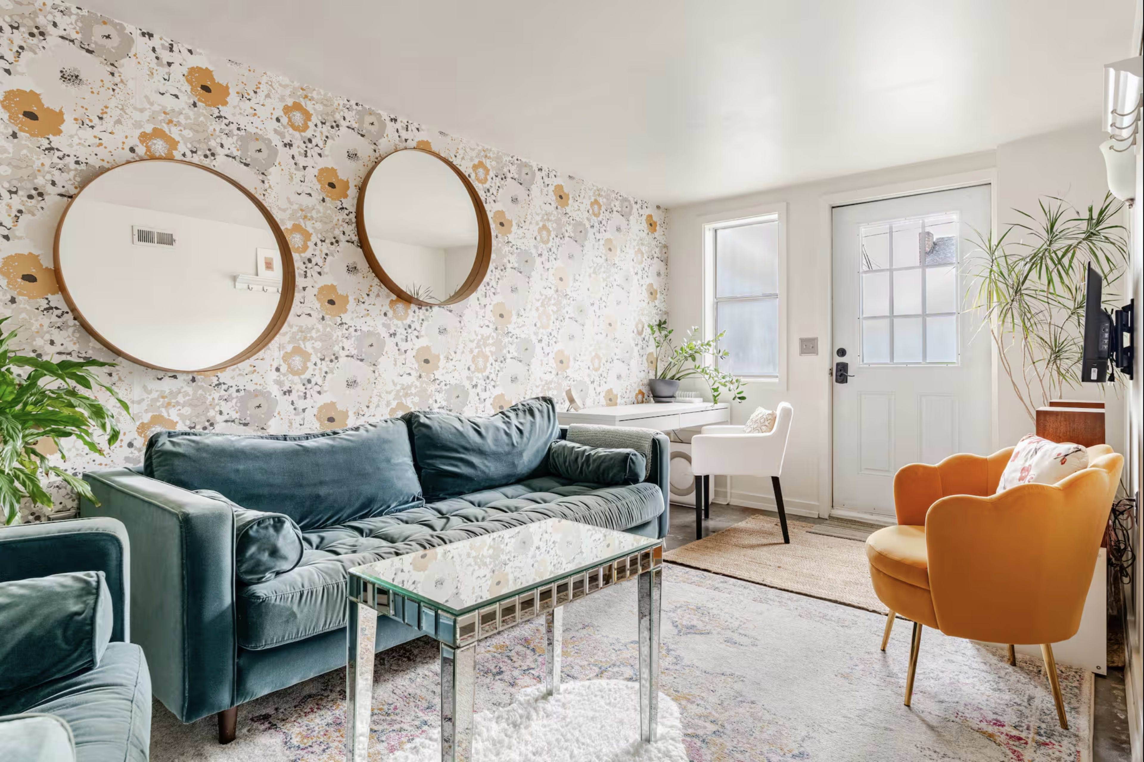 The image shows a cozy living room featuring a teal sofa, a glass coffee table, two round mirrors on the wall, and an orange armchair, with a floral wallpaper and a door leading outside.