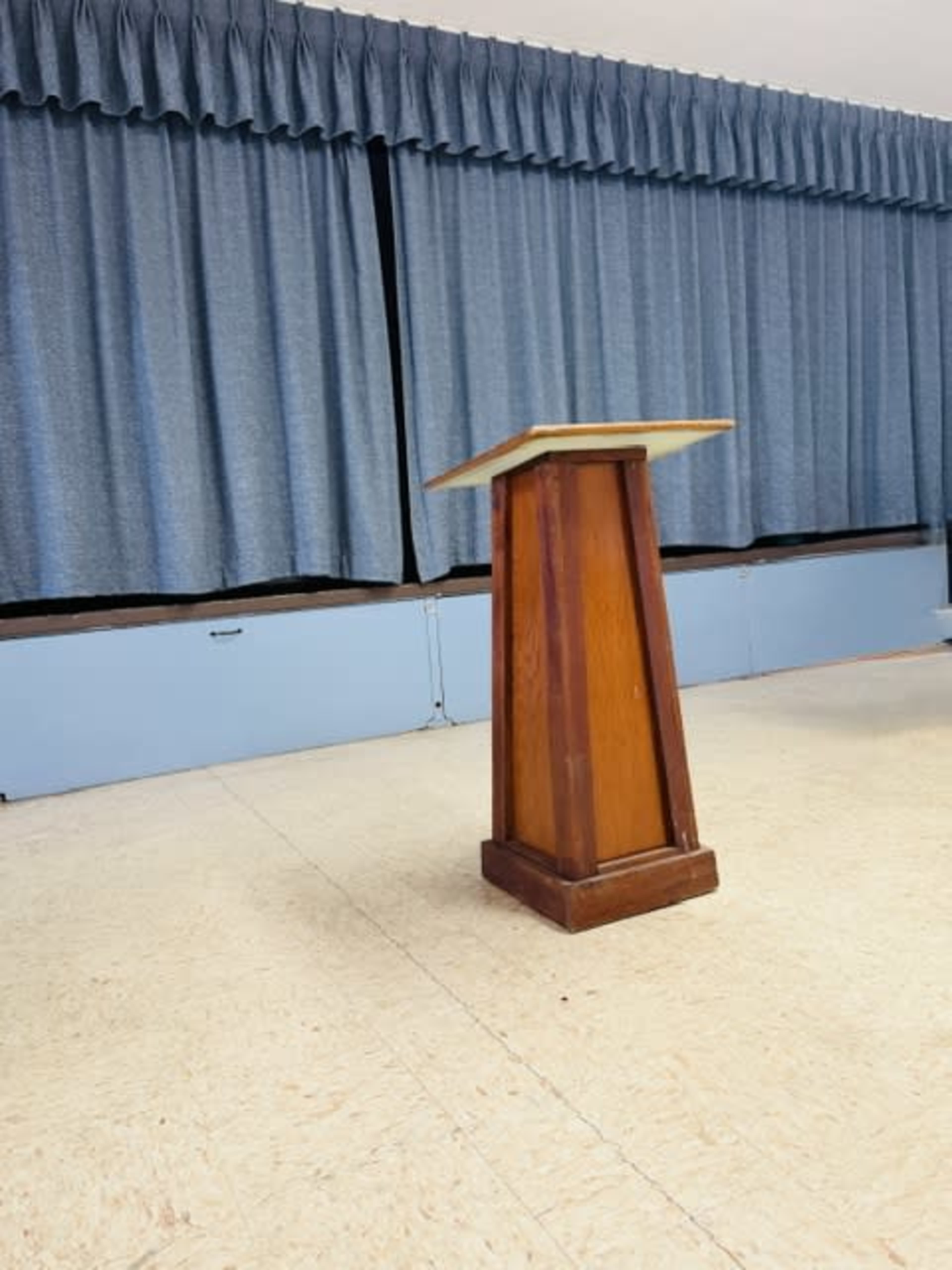 The image shows a wooden podium positioned on a light-colored floor, with blue curtains in the background.
