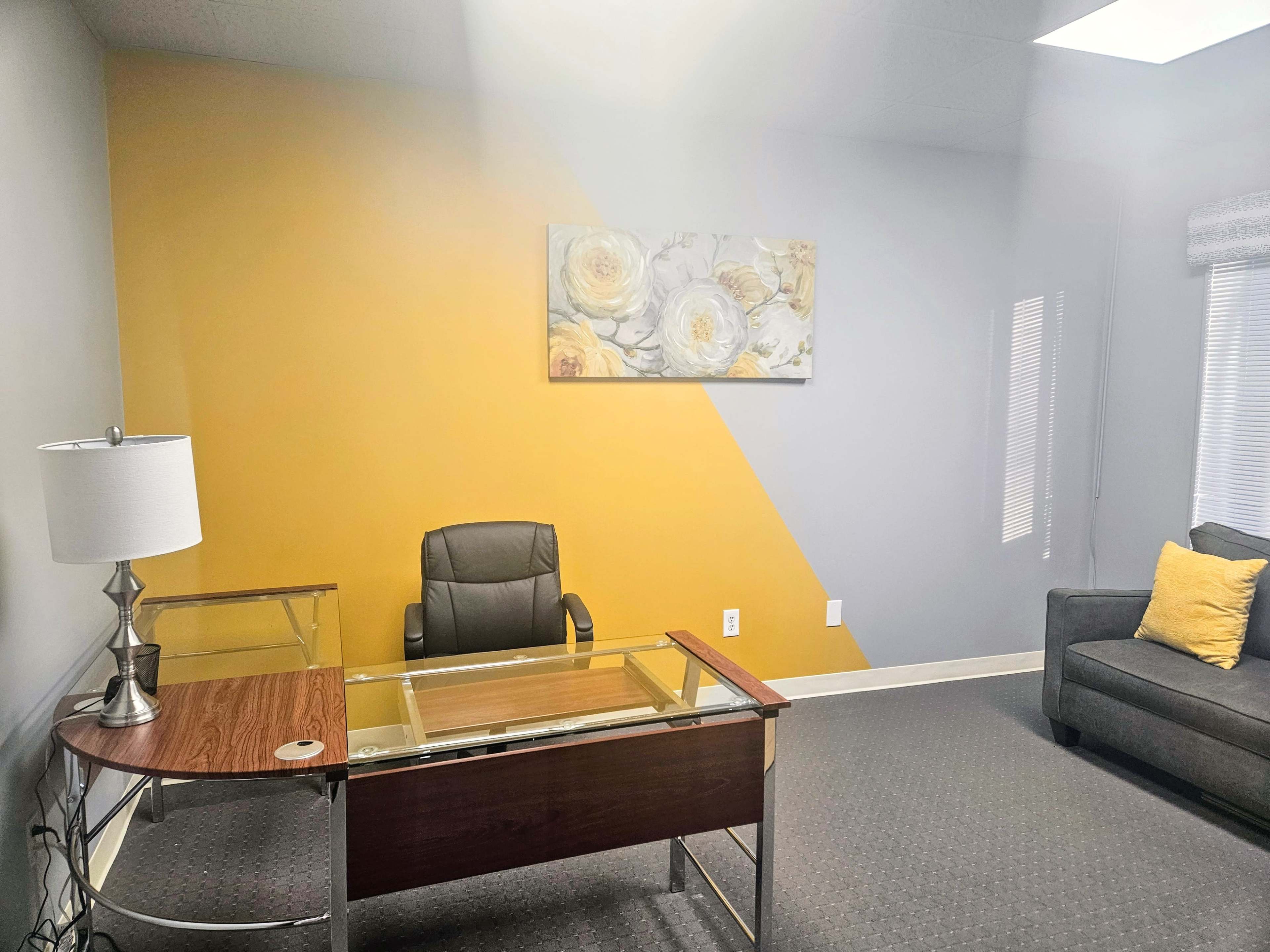 A well-lit office with a desk, an office chair, a sofa, and yellow and gray walls adorned with a floral painting.