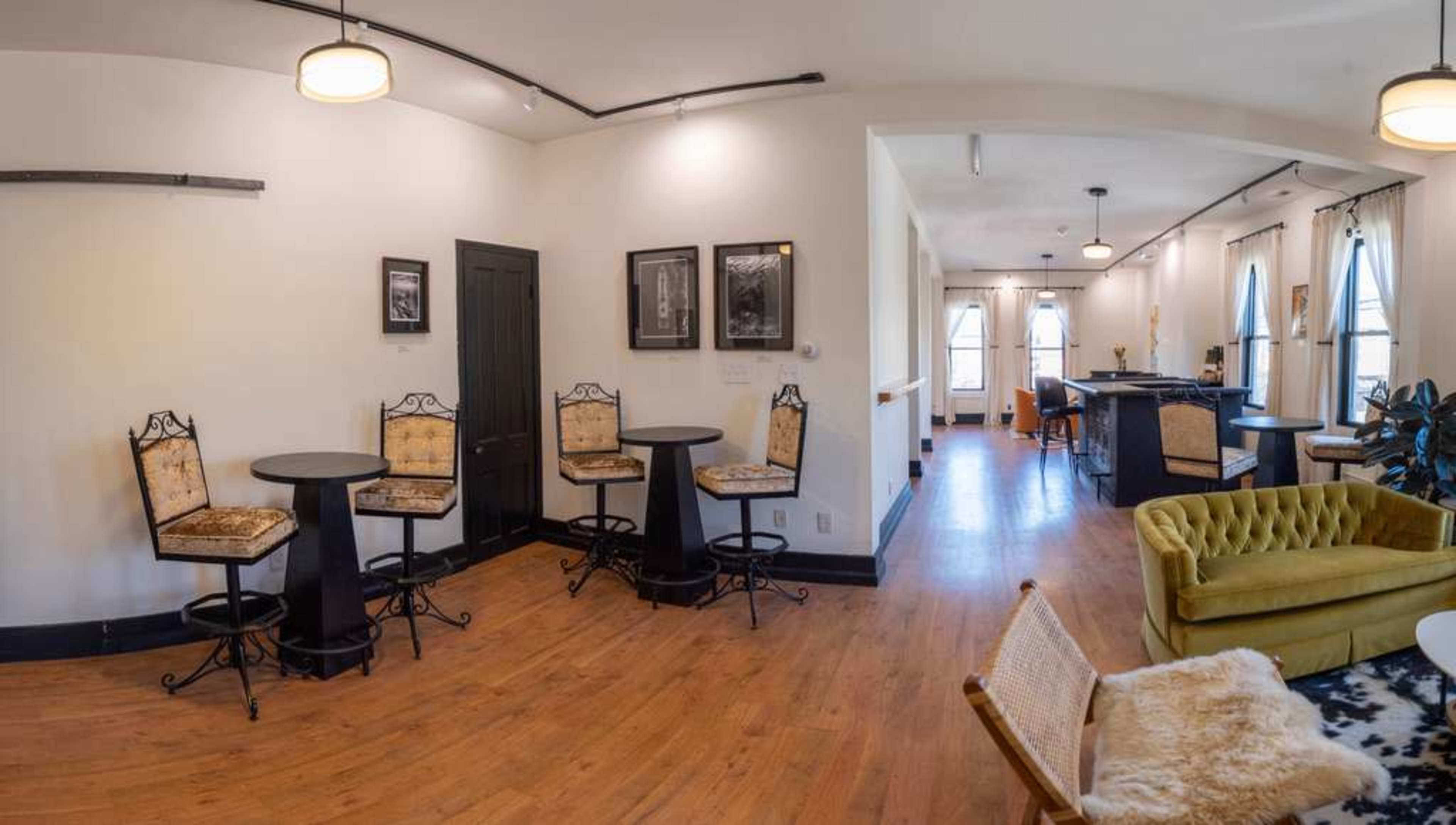 The image shows a spacious room with black and gold bar stools surrounding small tables, a green couch, and a well-lit area featuring large windows.