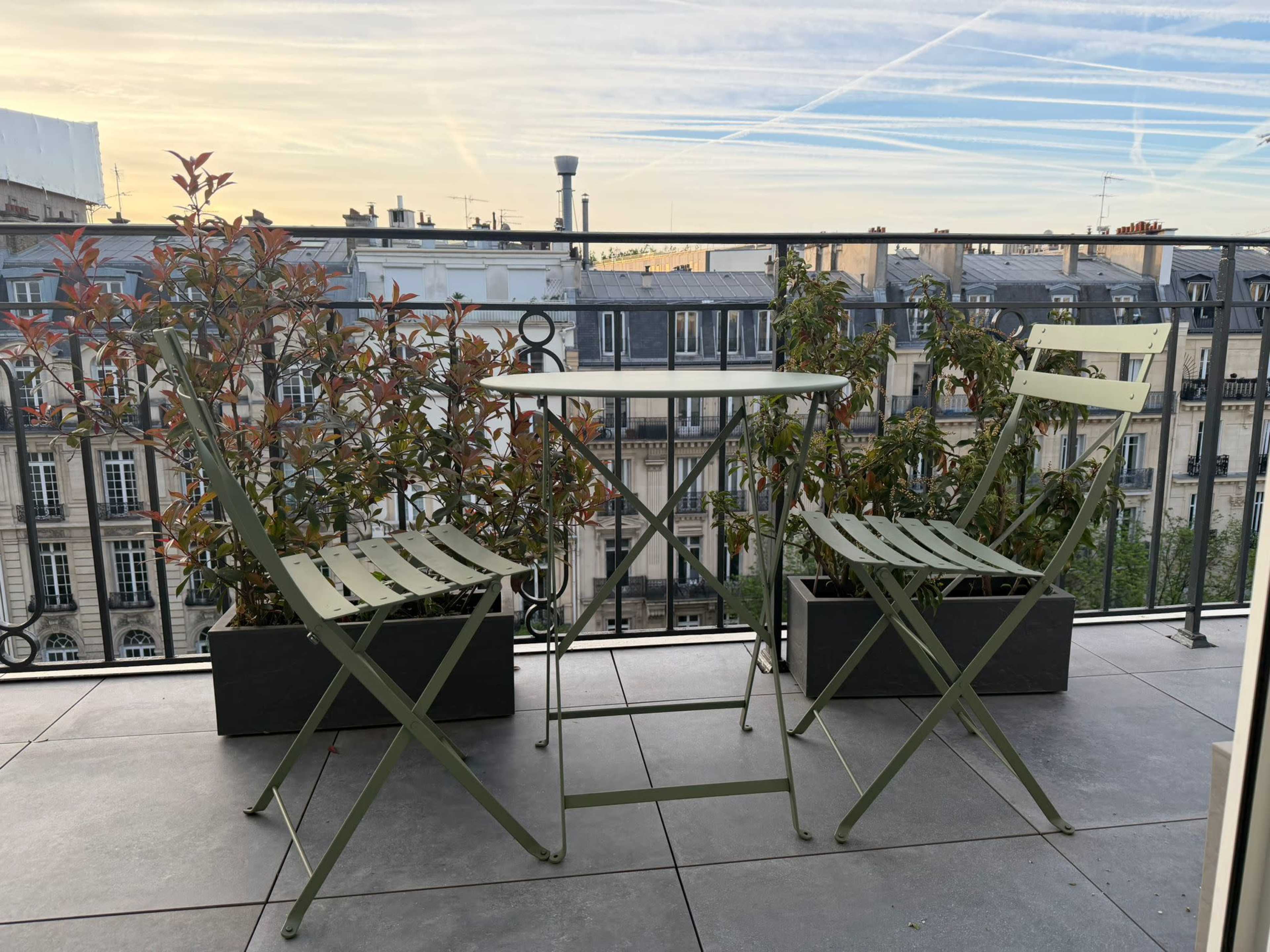 A small balcony features two green folding chairs, a table, and potted plants along the railing with a cityscape in the background.
