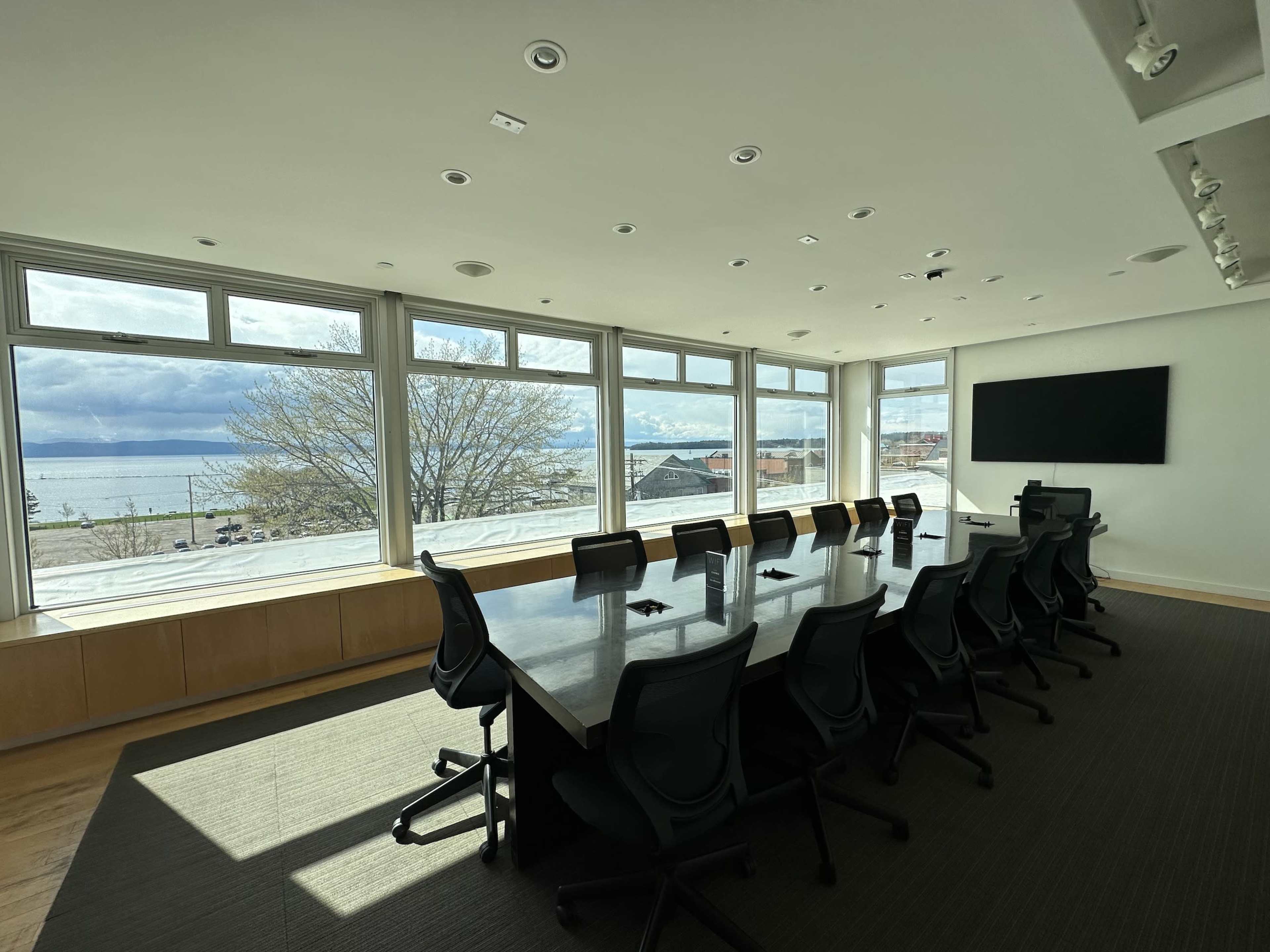A modern conference room features a long table surrounded by chairs, with large windows offering views of the nearby waterfront and landscape.