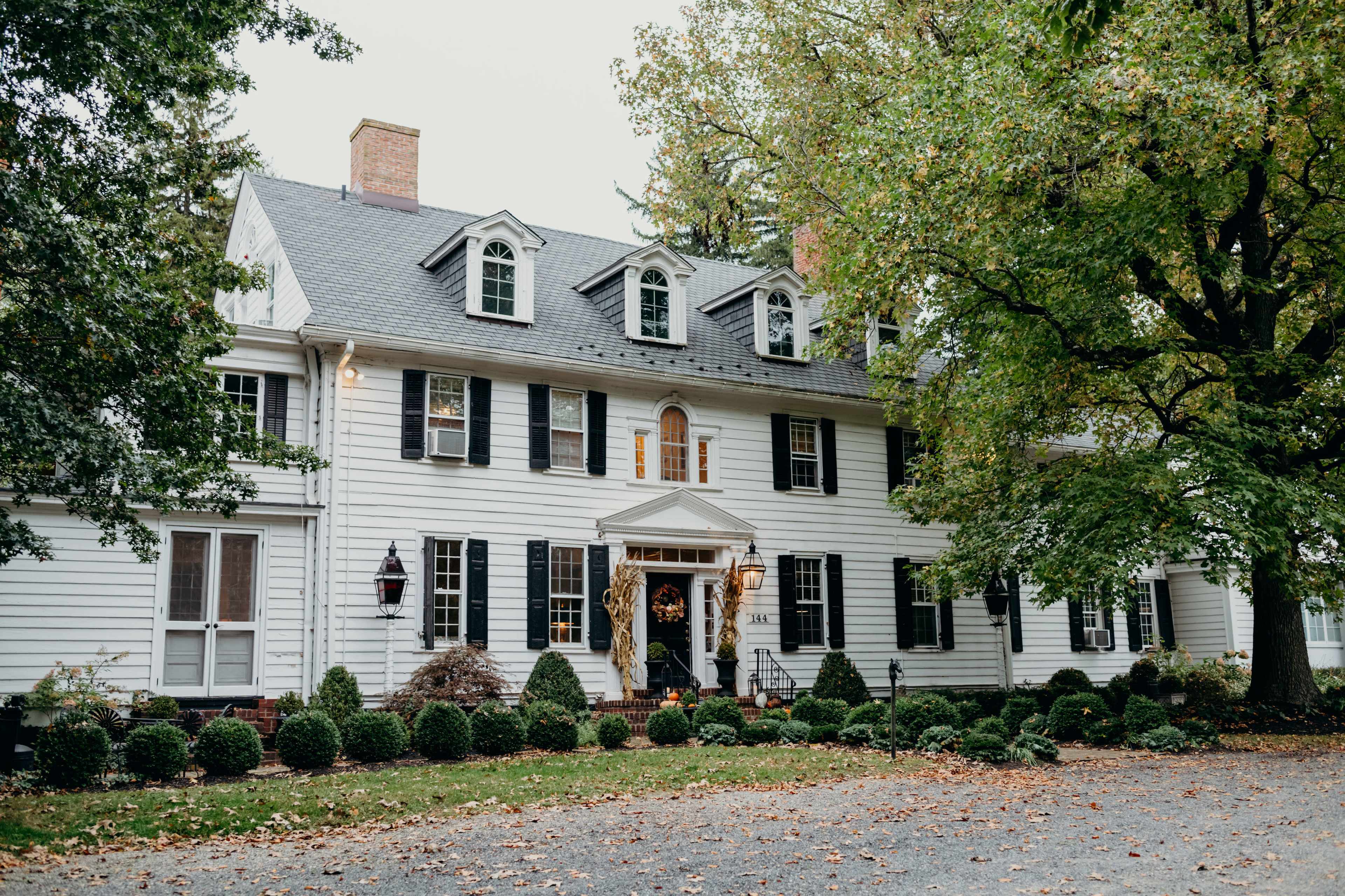 A large, classic white colonial house with black shutters and a centered front entrance is surrounded by neatly trimmed hedges and trees.