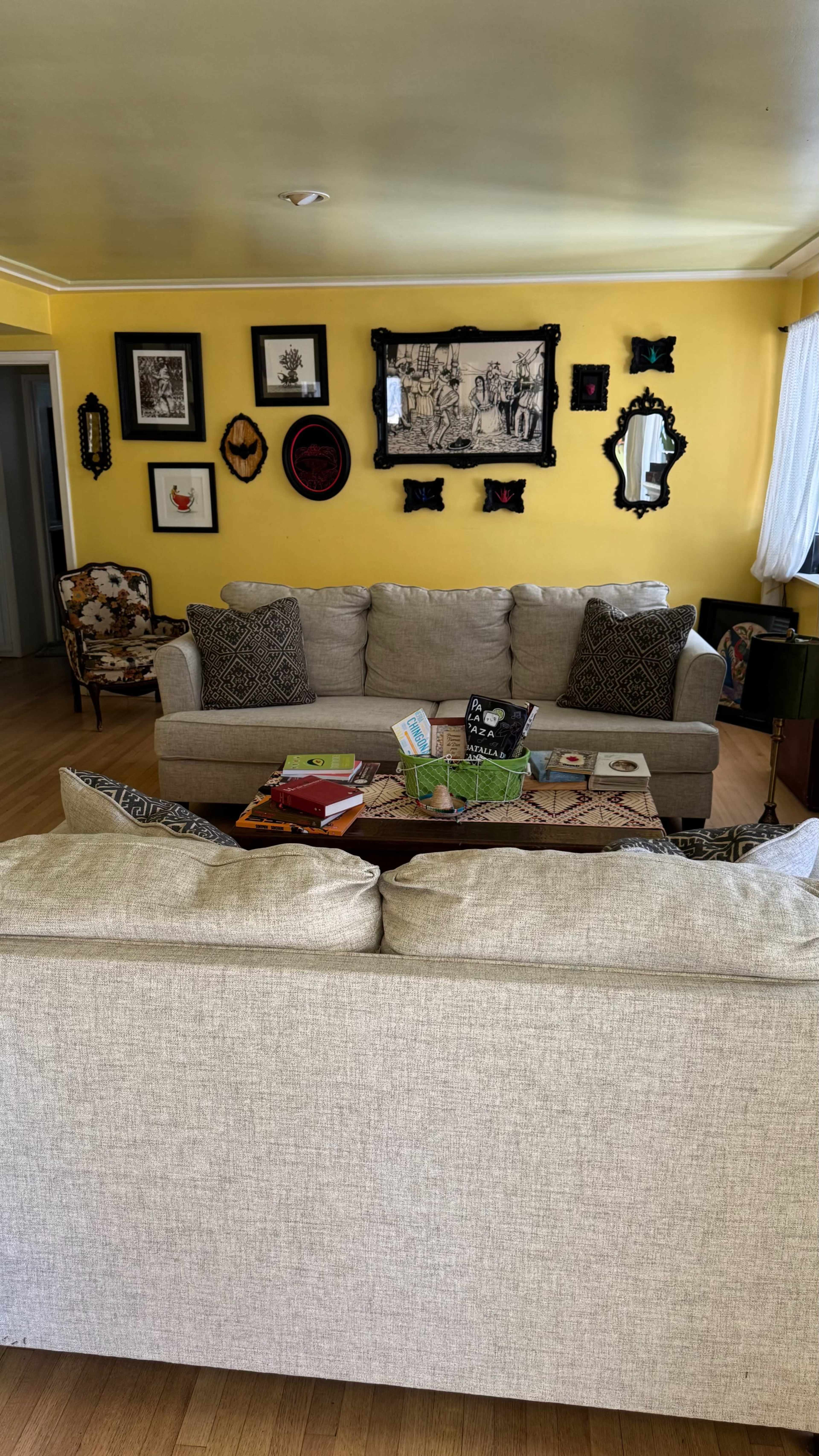 The living room features a light-colored sofa, a coffee table with various books, and an array of framed artwork and mirrors on the yellow walls.