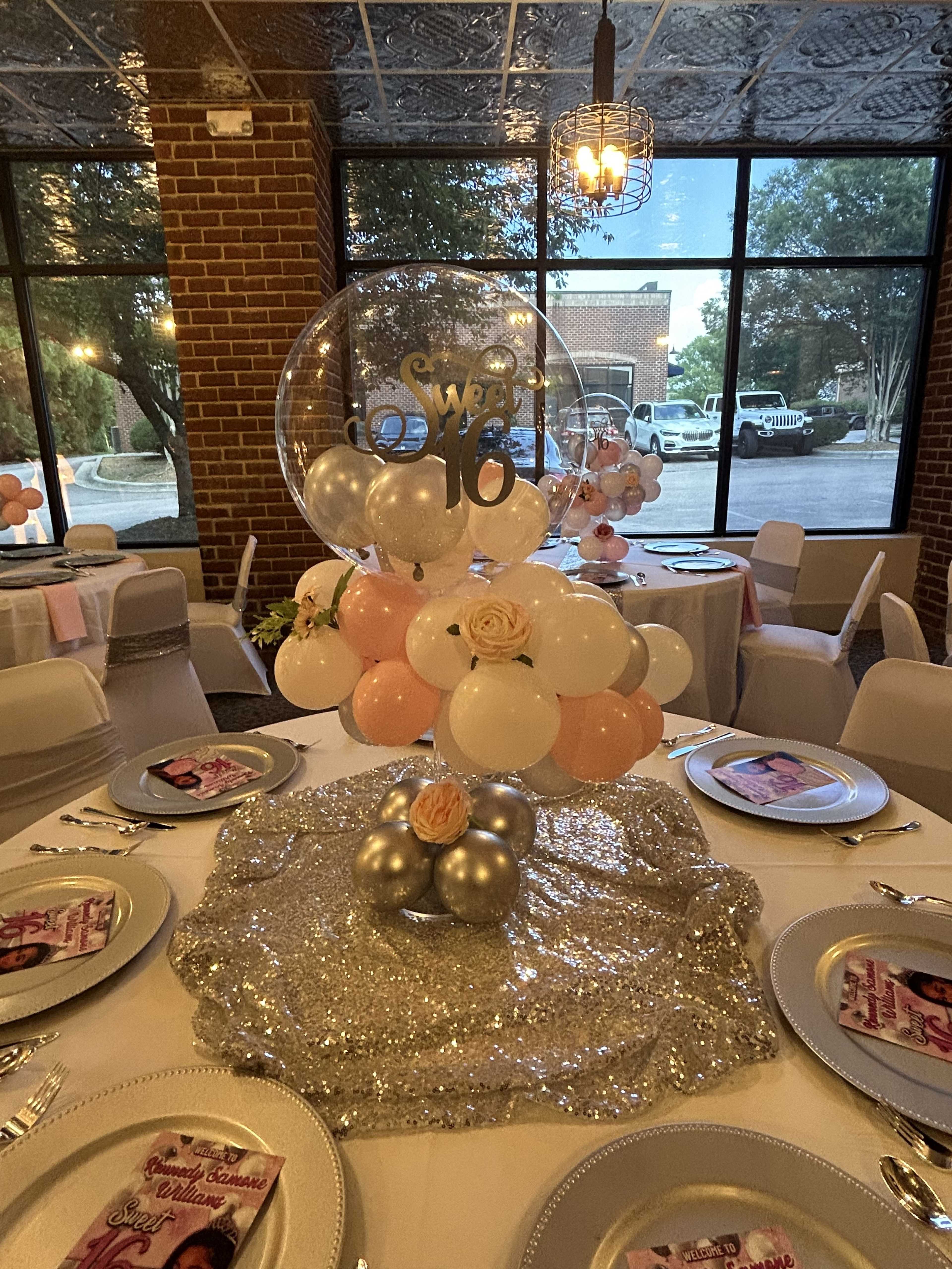 A table is set for a Sweet 16 celebration, featuring a decorative centerpiece of balloons and a floral accent on a sparkly tablecloth.