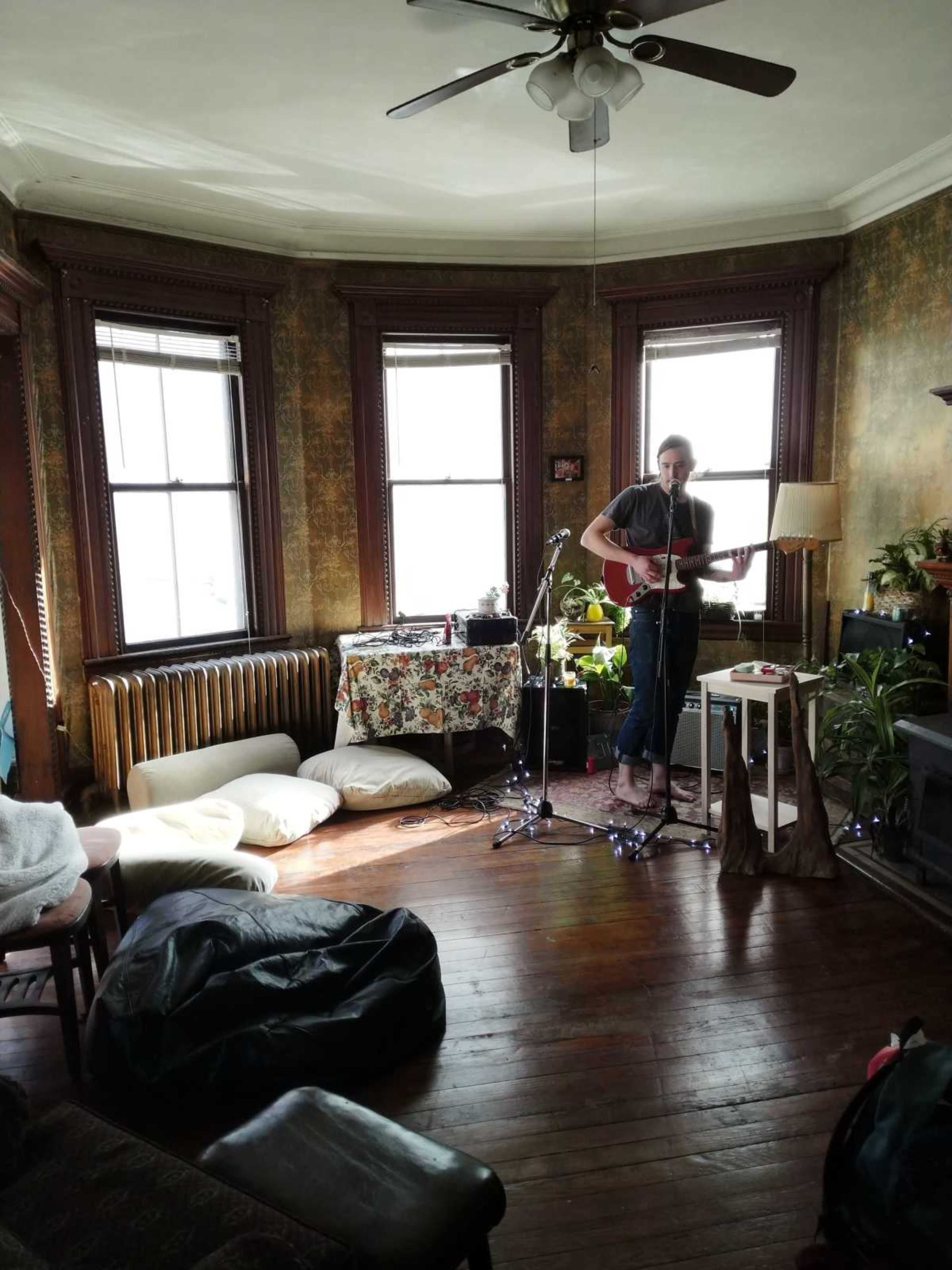 A musician stands playing guitar in a cozy room with vintage decor, surrounded by plants and various seating options.