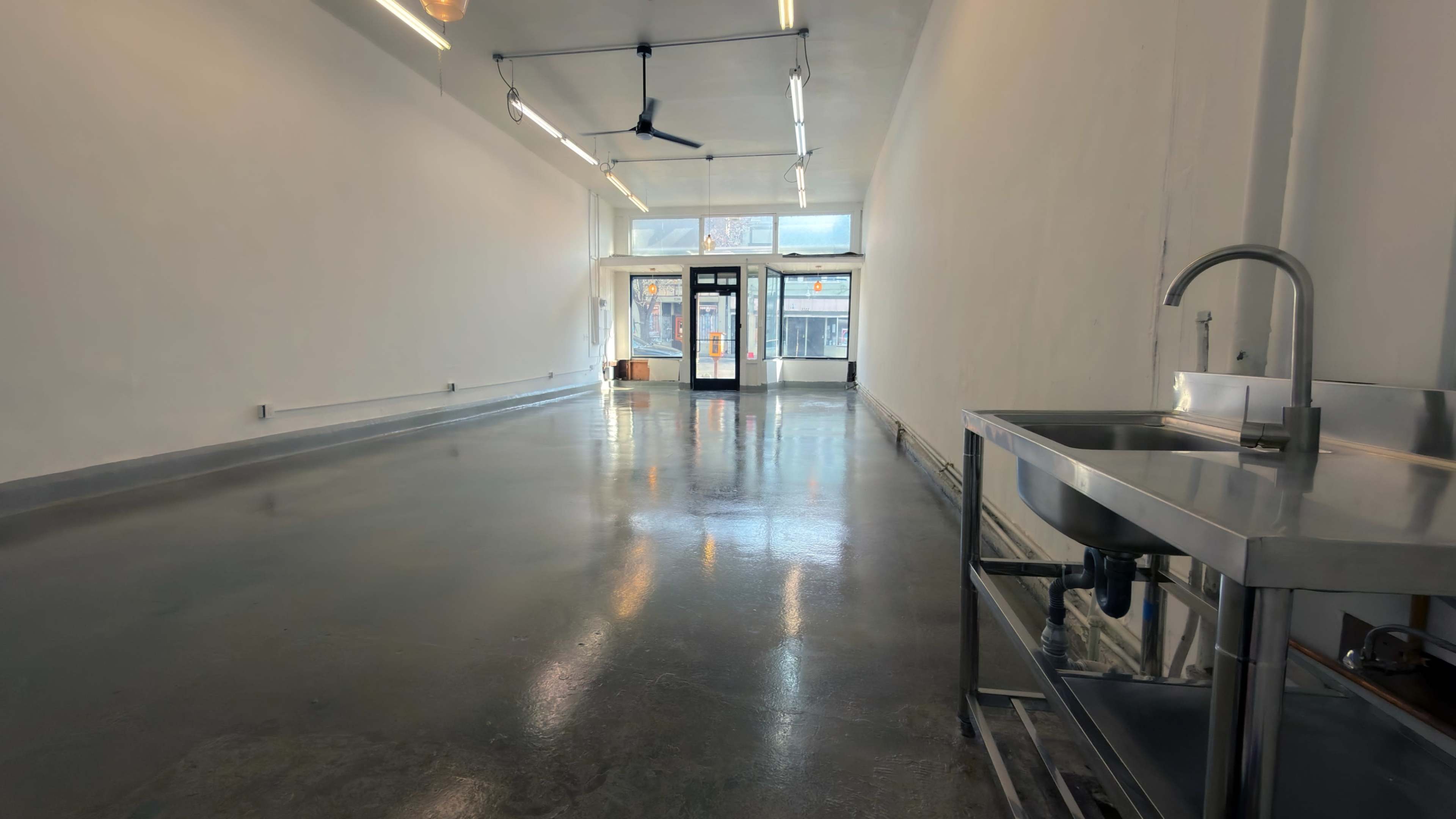 The image shows a vacant commercial space with polished concrete flooring, fluorescent lighting, a ceiling fan, and a stainless steel sink on a rolling cart near the left side.