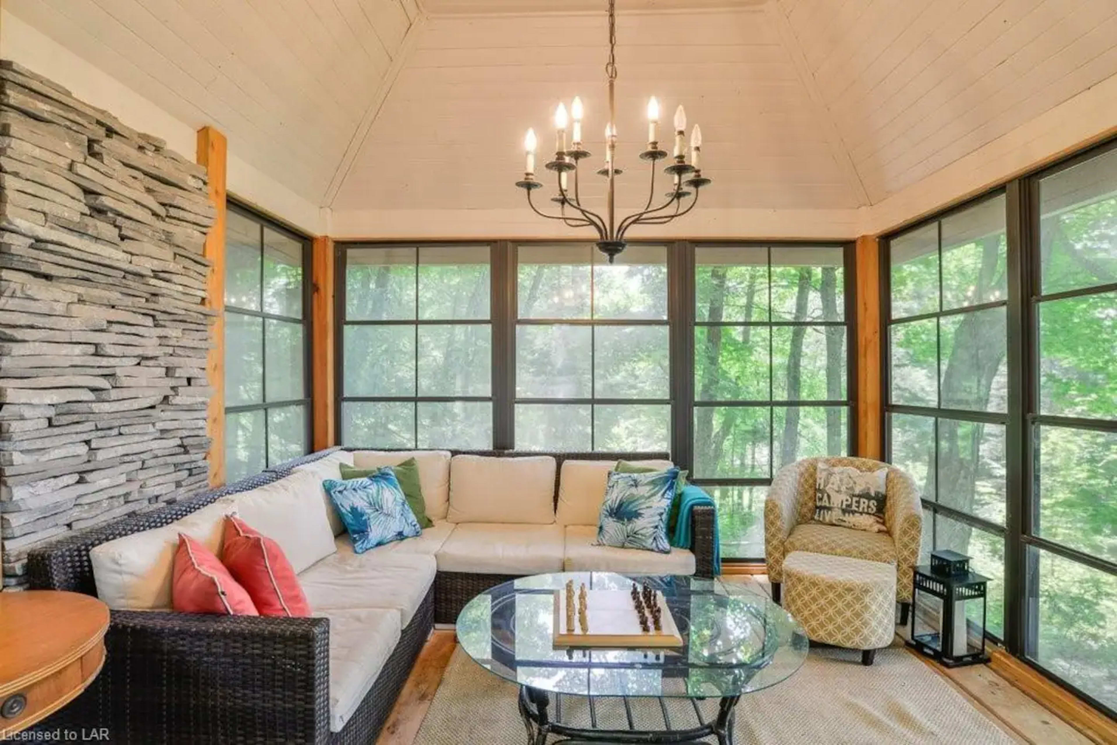 A sunroom features a sectional sofa, a glass coffee table with a chess set, and a chandelier, surrounded by large windows providing views of the outdoors.