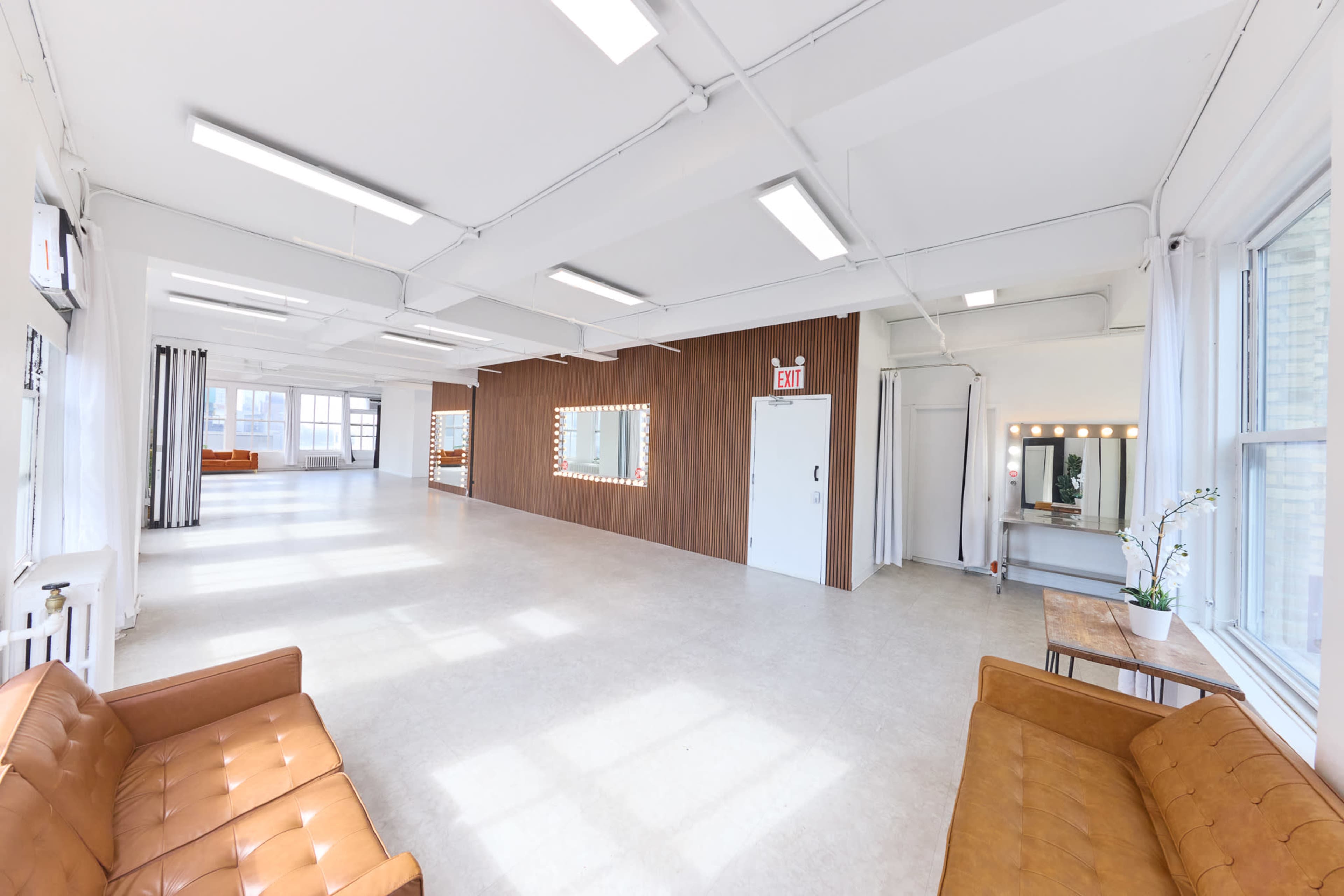 The image depicts a spacious, well-lit interior room with beige flooring, white walls, and a wooden accent wall featuring a mirror.