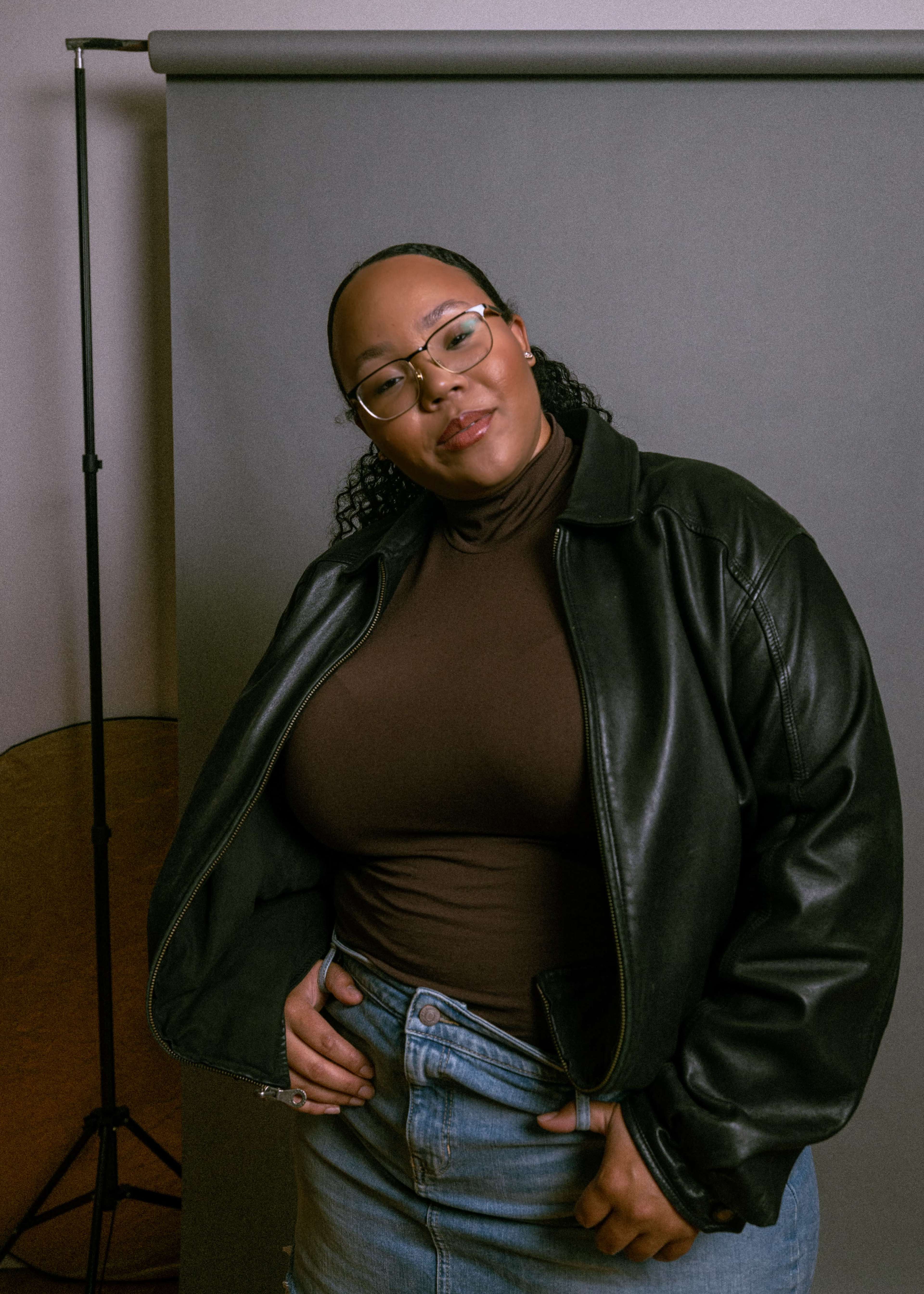 A woman wearing a brown turtleneck and a black leather jacket stands in front of a gray backdrop.