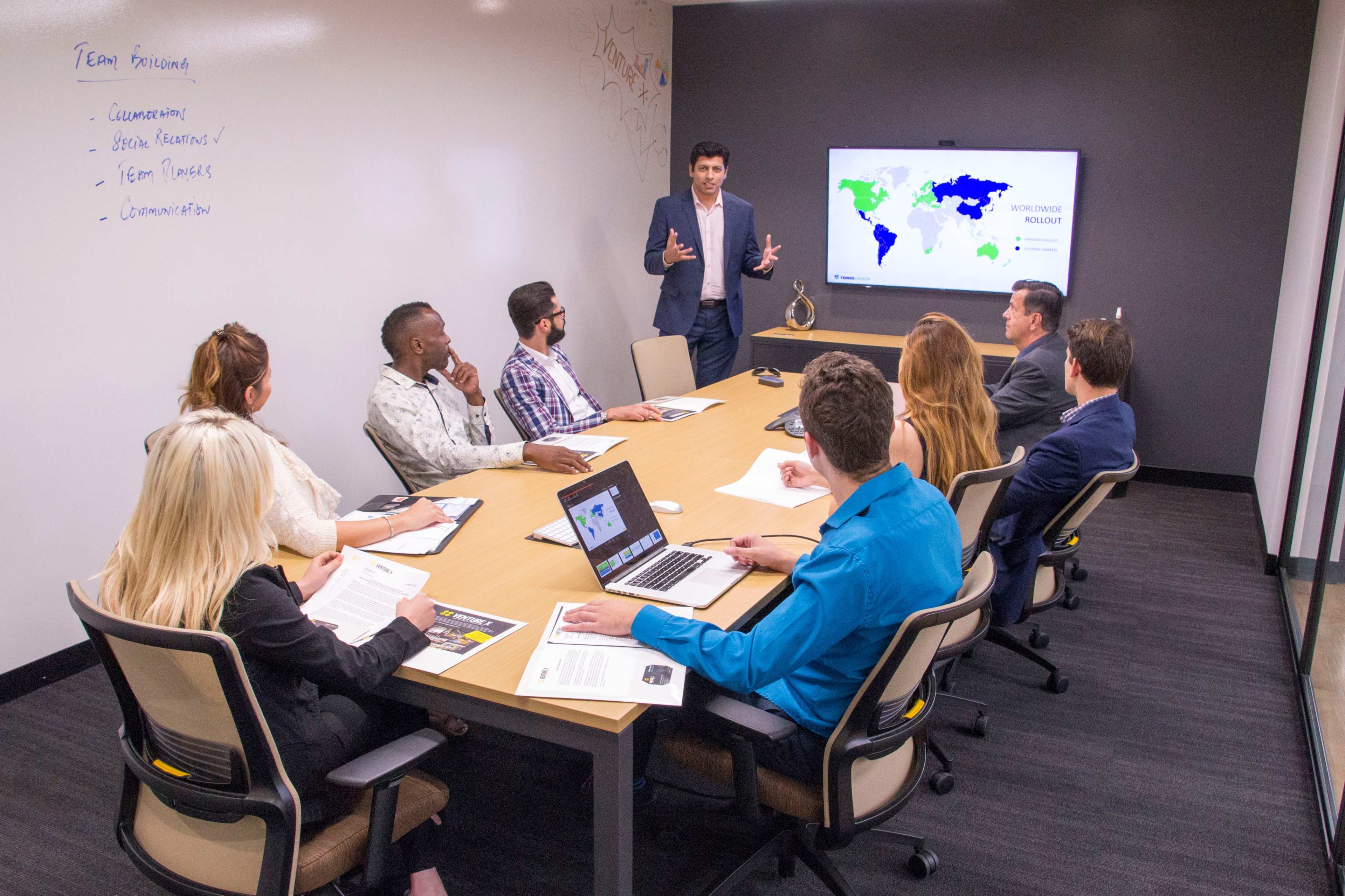 A diverse group of professionals is gathered around a conference table in a meeting room, with a speaker presenting a world map on a screen.